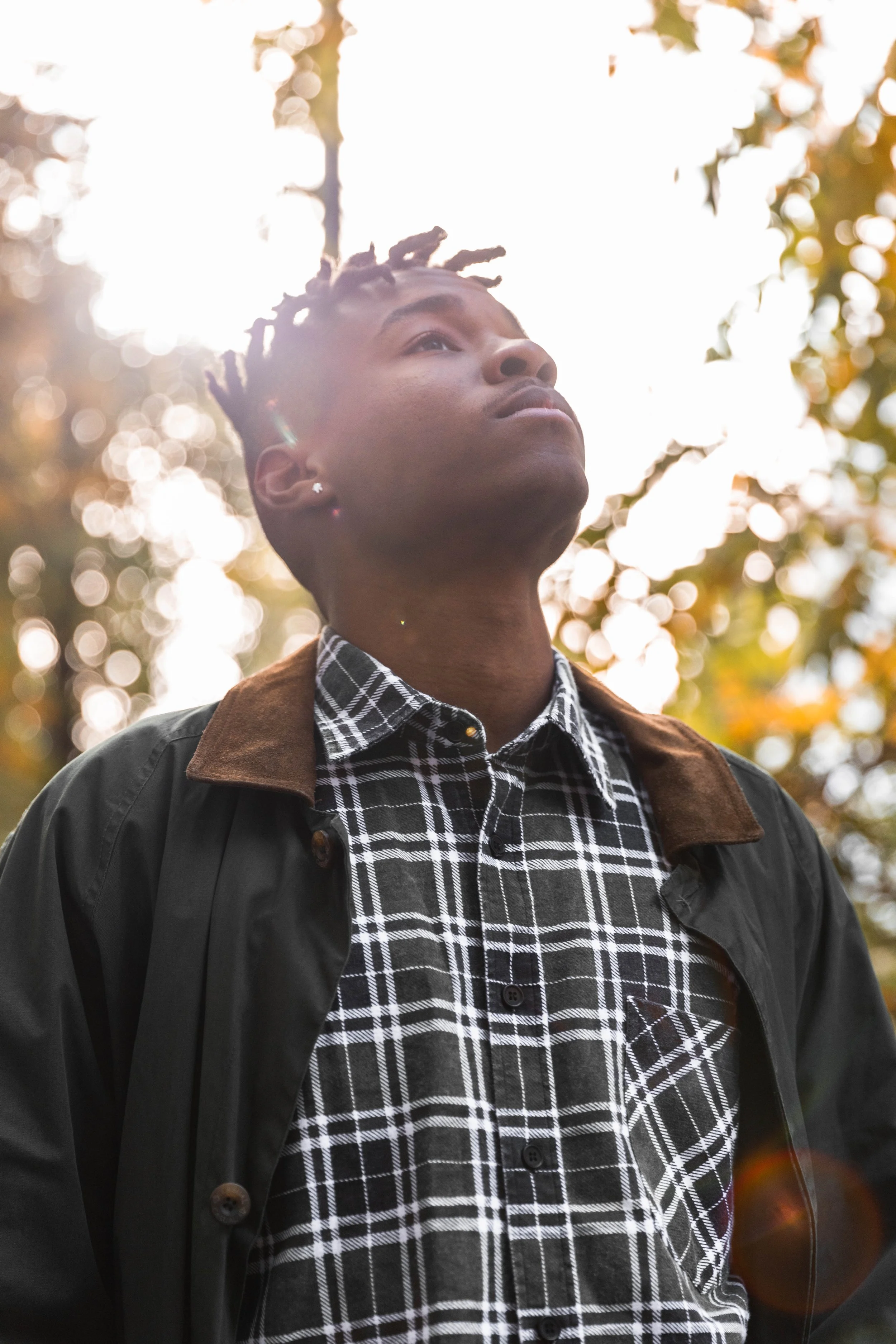 Jeune homme regardant vers le haut, en plein air, avec lumière du soleil en arrière-plan, portant une chemise à carreaux noir et blanc et une veste foncée.