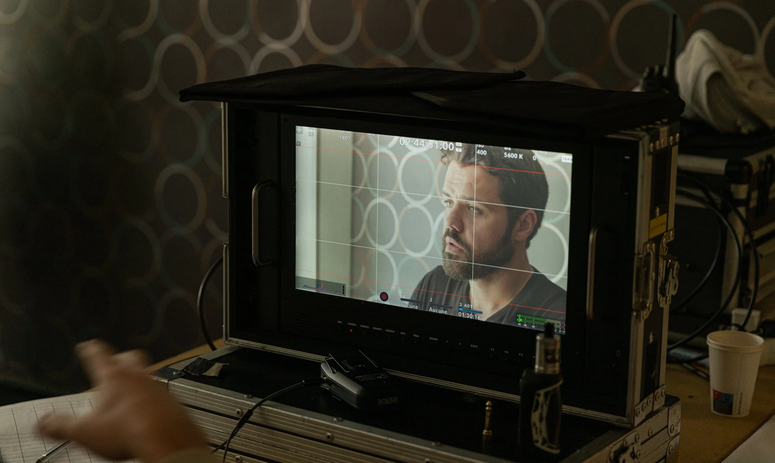 Un homme avec une barbe regarde une caméra sur un écran, en train de filmer une scène. Derrière, il y a un mur avec un motif de cercles.