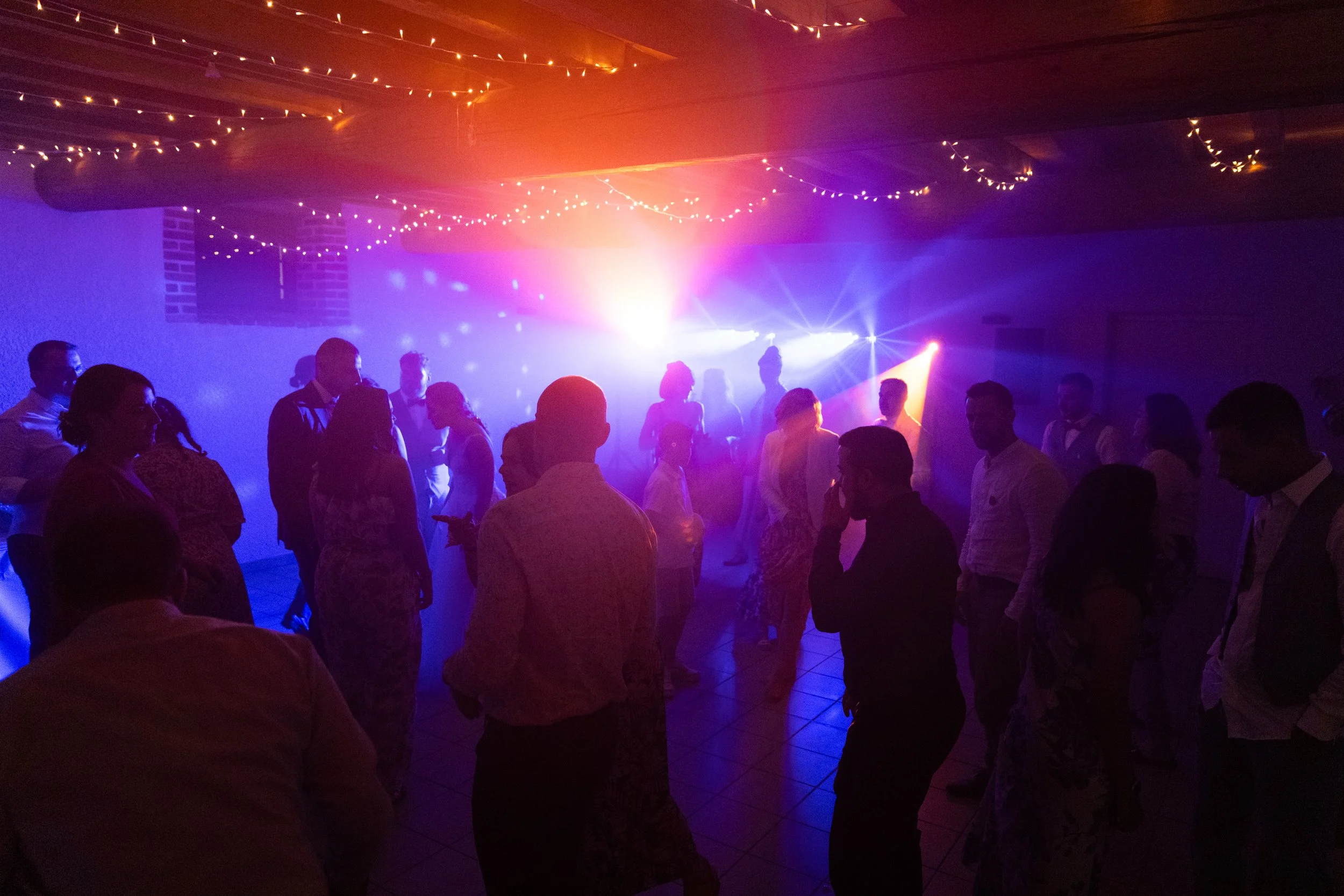 Groupe de personnes dansant dans une salle éclairée avec des lumières colorées et des guirlandes lumineuses suspendues au plafond.