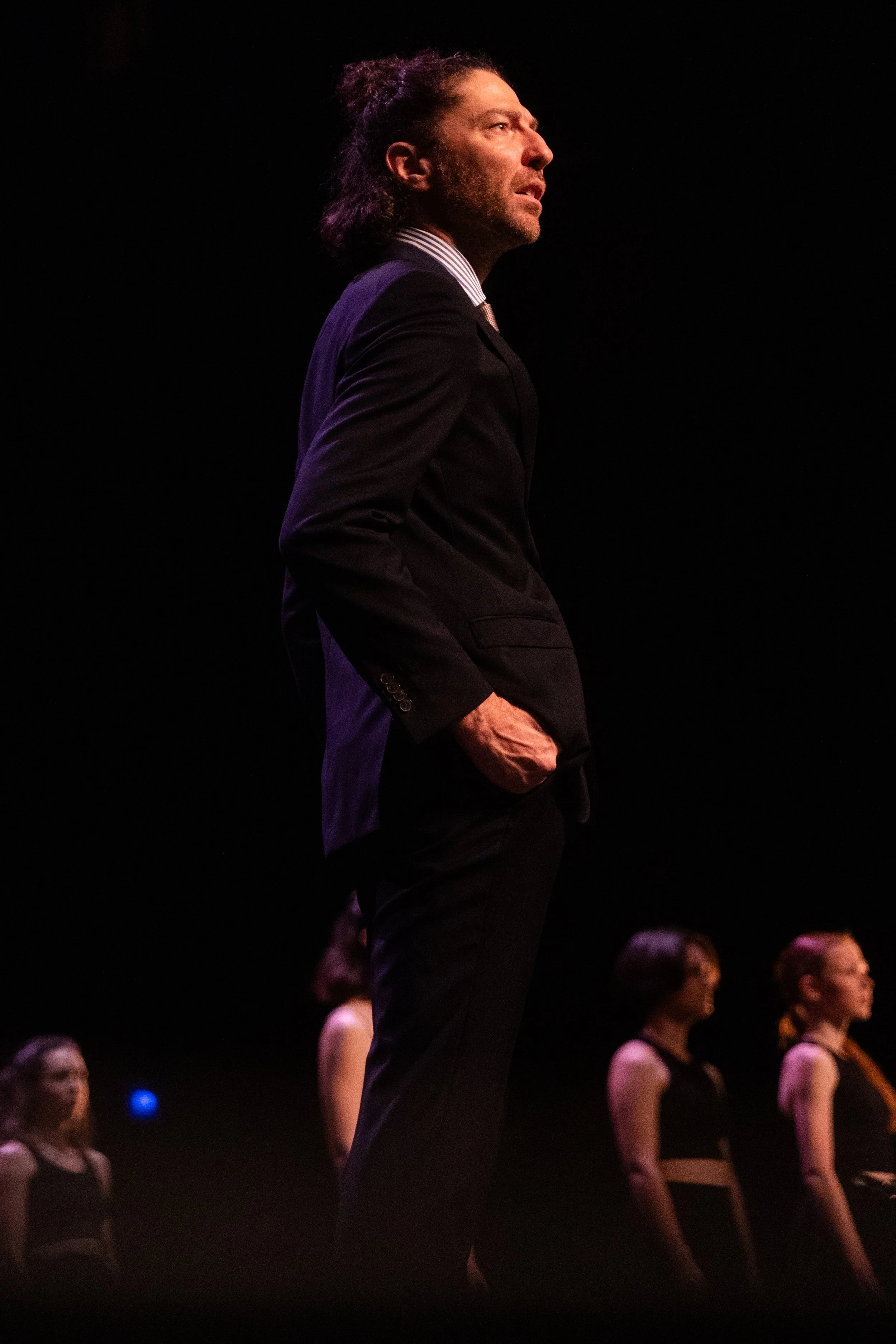 Un homme en costume noir se tient debout avec les mains dans les poches, entouré de plusieurs femmes dans un espace sombre, probablement une scène de spectacle ou une répétition.