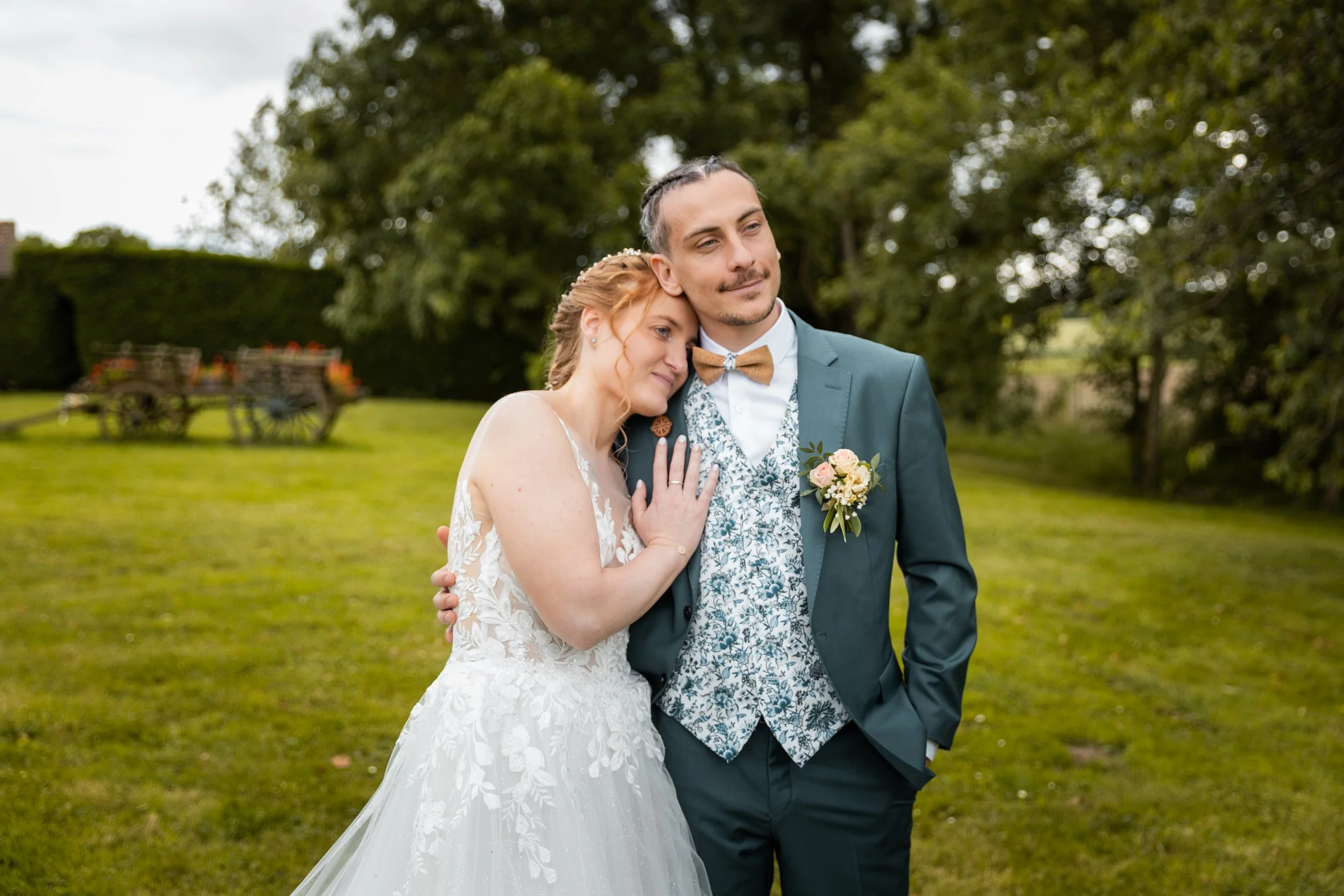 Un couple en tenue de mariage pose dans un jardin, l'homme porte un costume vert avec un nœud papillon en bois et la femme porte une robe de mariée en dentelle, elle se repose sa tête sur l'épaule de l'homme.