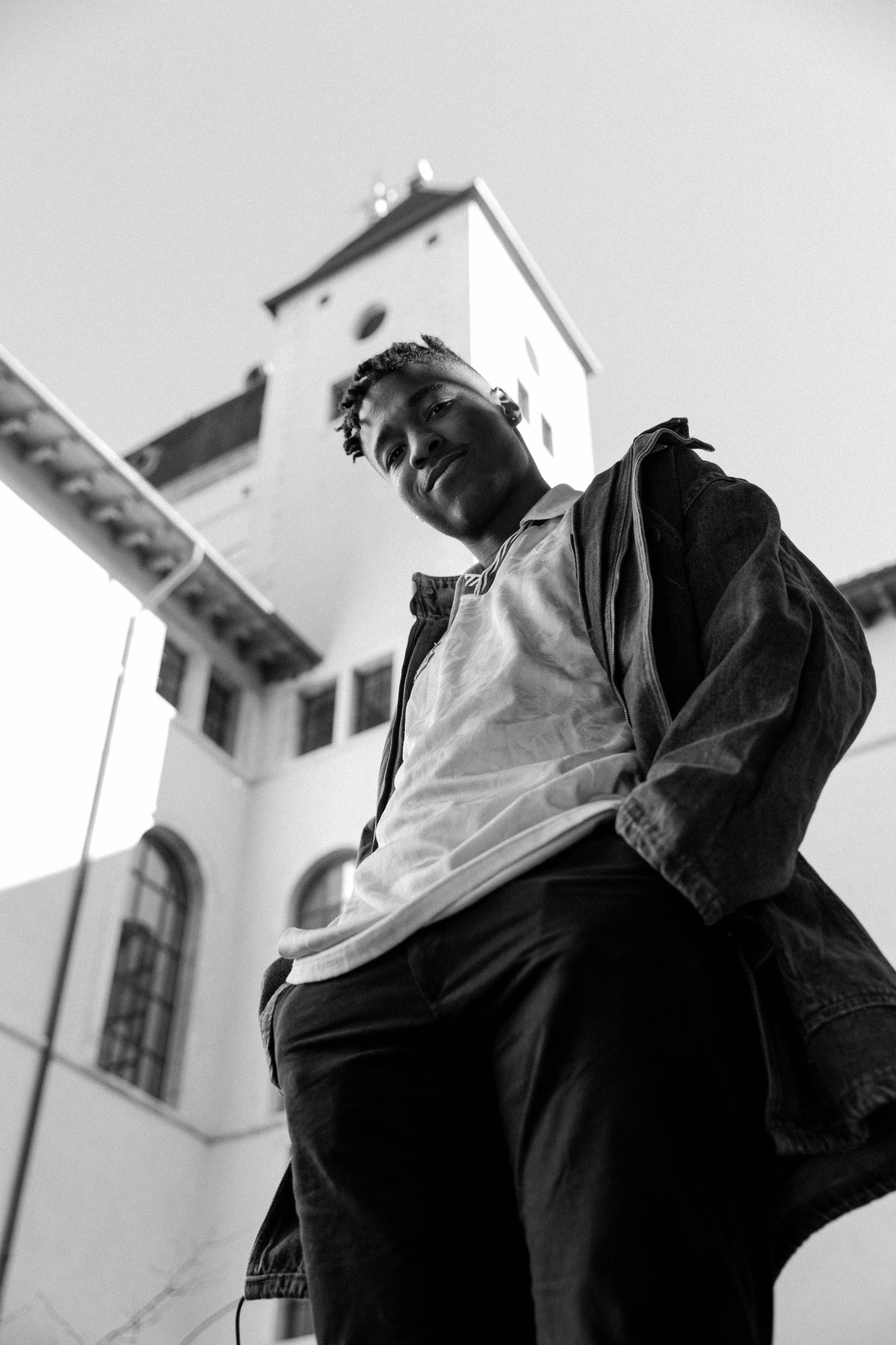 Un jeune homme pose devant un bâtiment avec une tour, vue en contre-plongée, en noir et blanc.