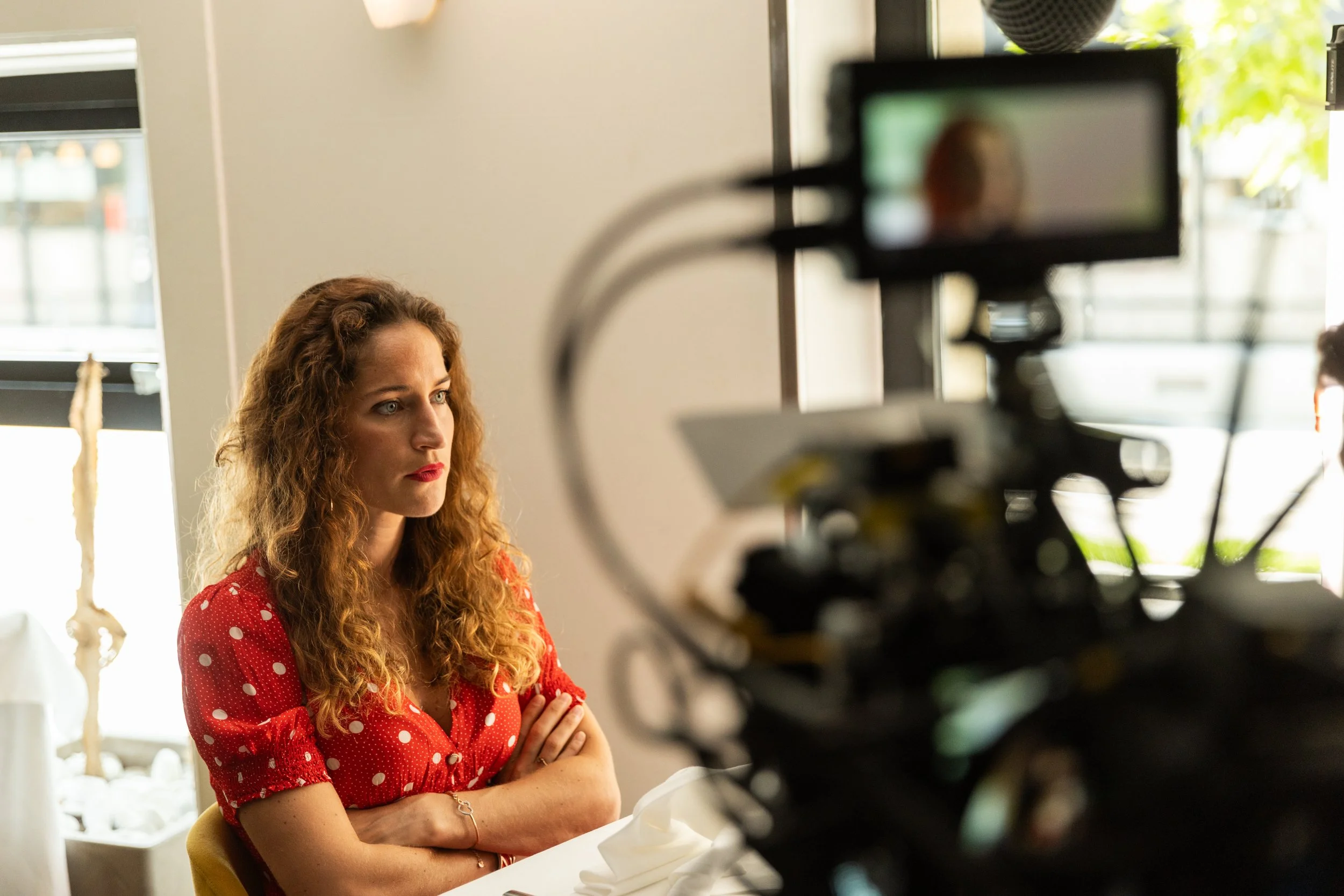 Une femme aux cheveux bouclés, portant une robe rouge à pois blancs, assise dans une pièce avec une caméra de tournage en premier plan.