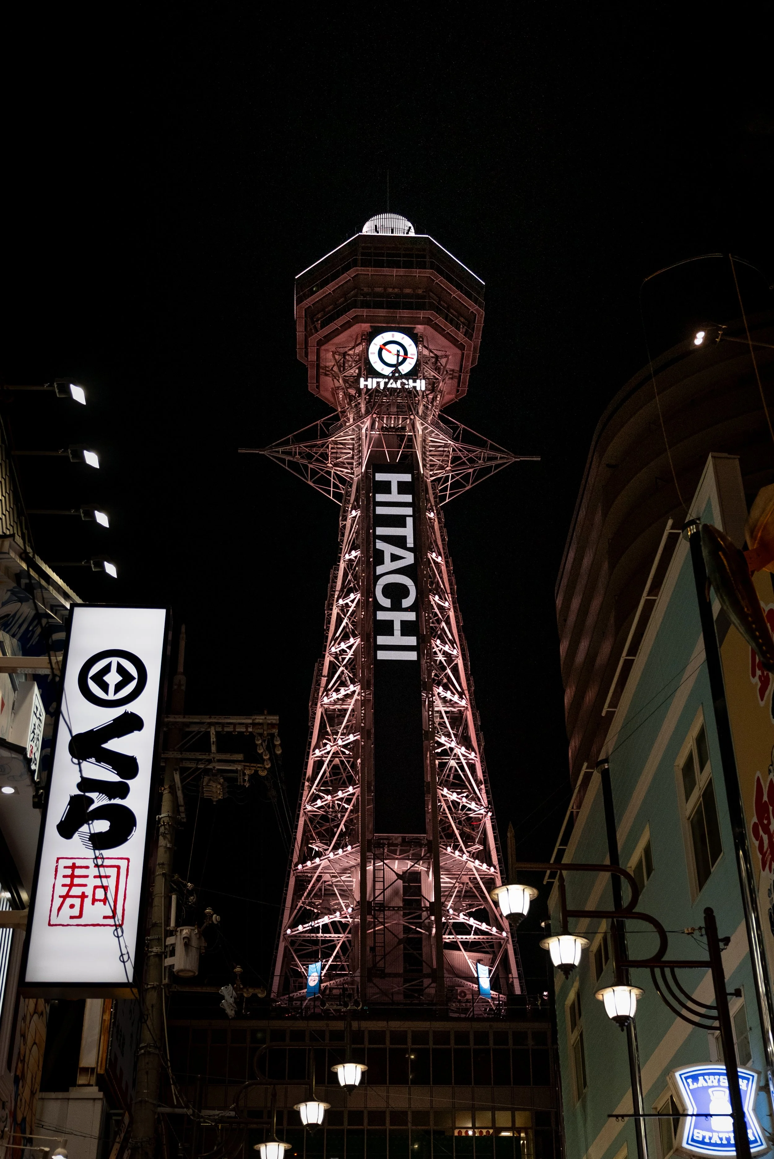 Tour de télévision Hitachi illuminée la nuit, vue en contre-plongée avec des enseignes lumineuses environnantes.