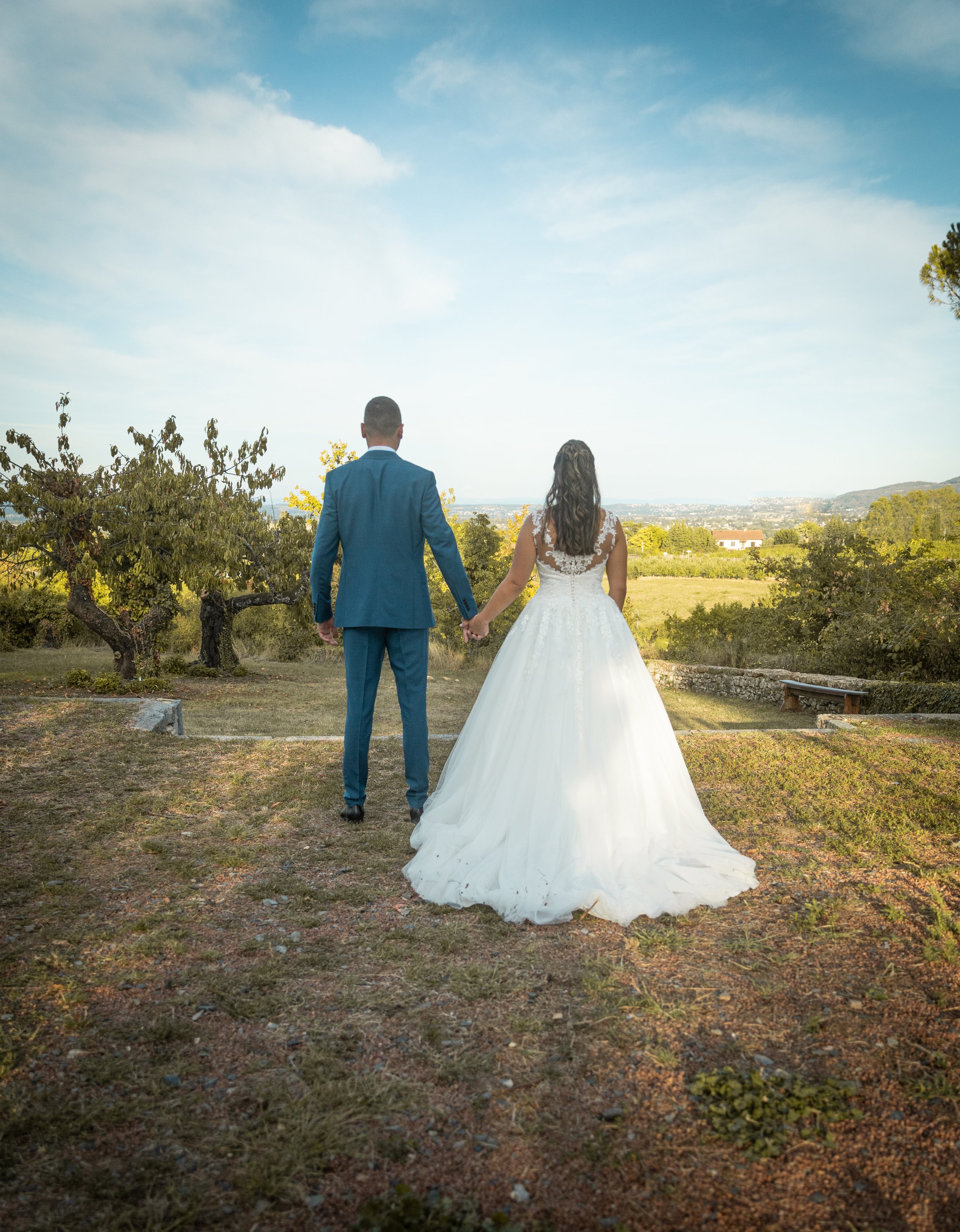 Un couple de mariés marche main dans la main dans un champ en plein air, avec un ciel bleu et des arbres en arrière-plan.