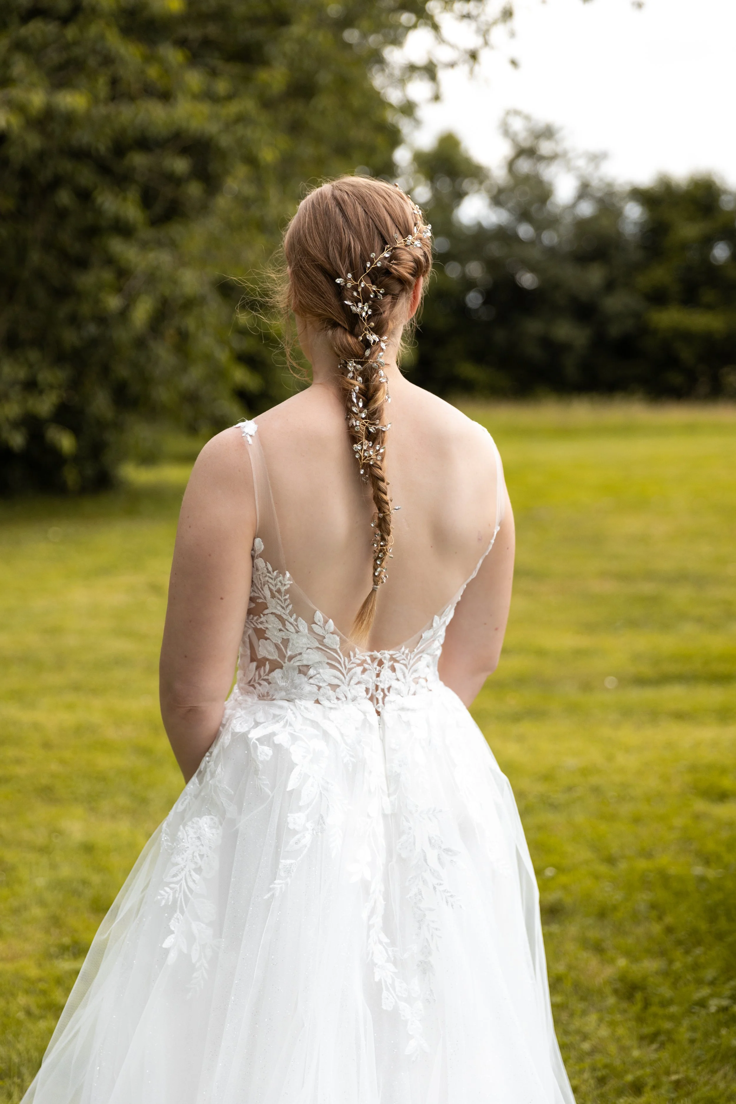 Une femme en robe de mariage blanche avec un dos en V, coiffée d'une tresse ornée de décorations florales, dans un parc vert.