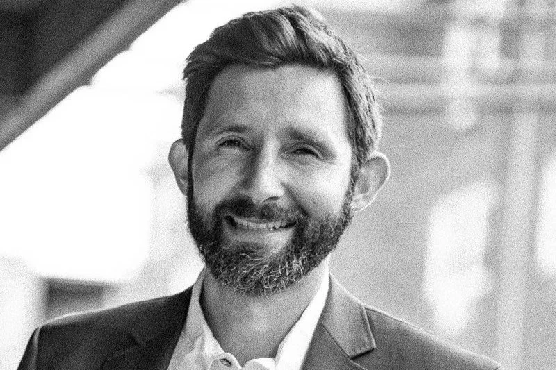 Black and white portrait of Michael with a beard and short hair, smiling, wearing a suit, in front of a background with blurred architectural elements.