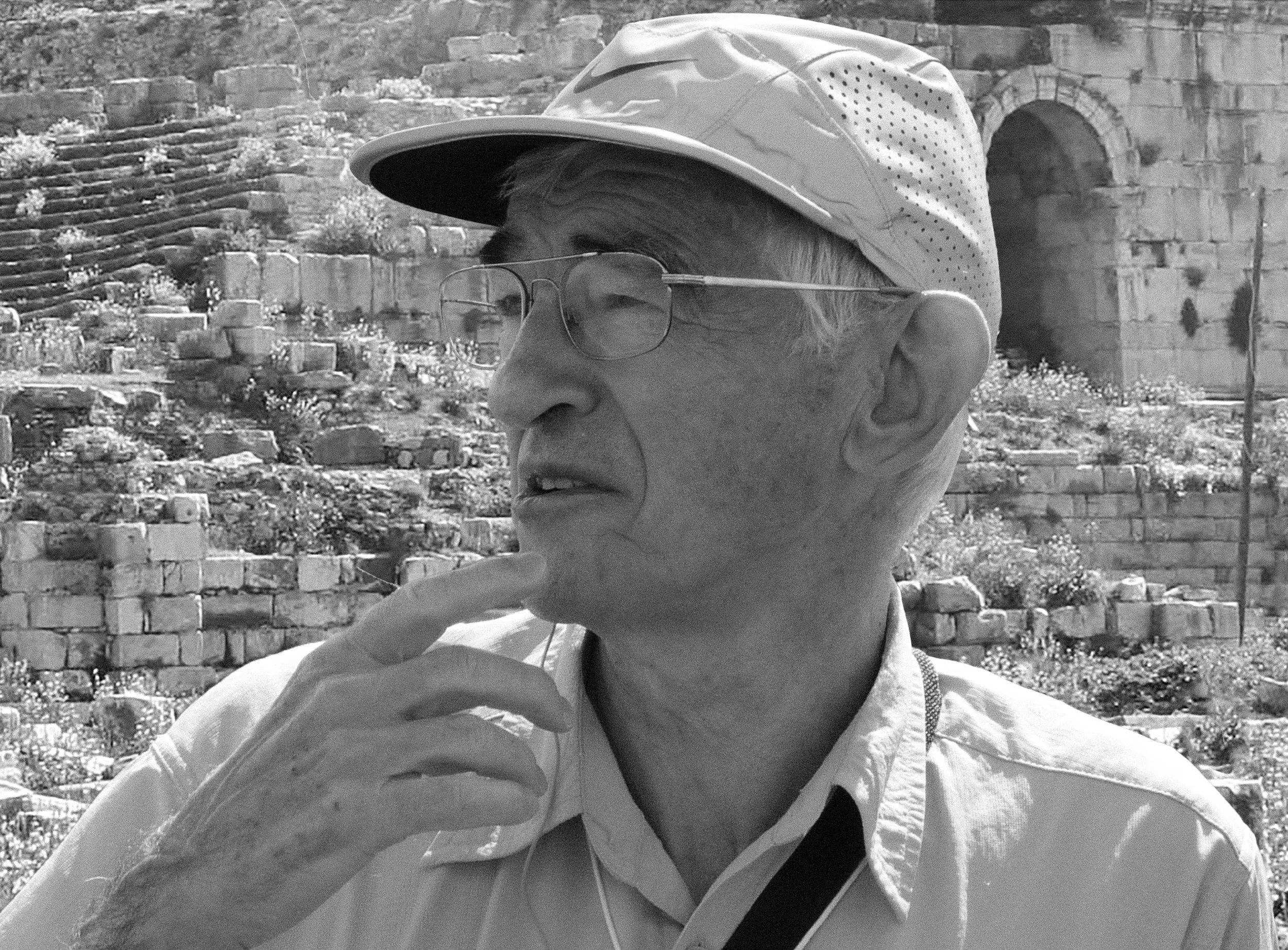 A black and white photo of John Dominic wearing a cap and glasses, with his hand touching his chin in thought in front of a stone structure resembling ruins.