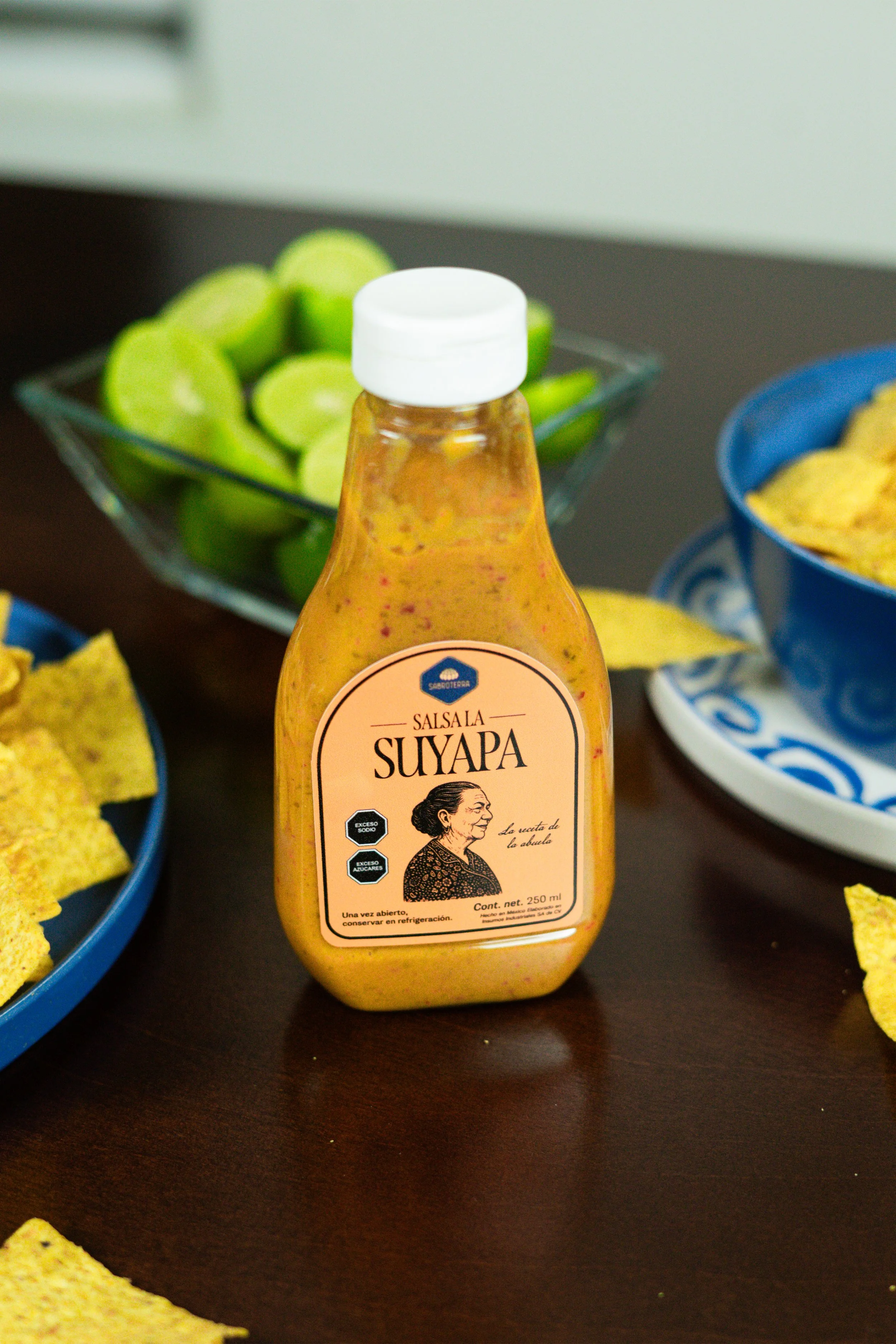 Botella de salsa SUYAPA en una mesa, rodeada de totopos y limones cortados en un plato de cristal.