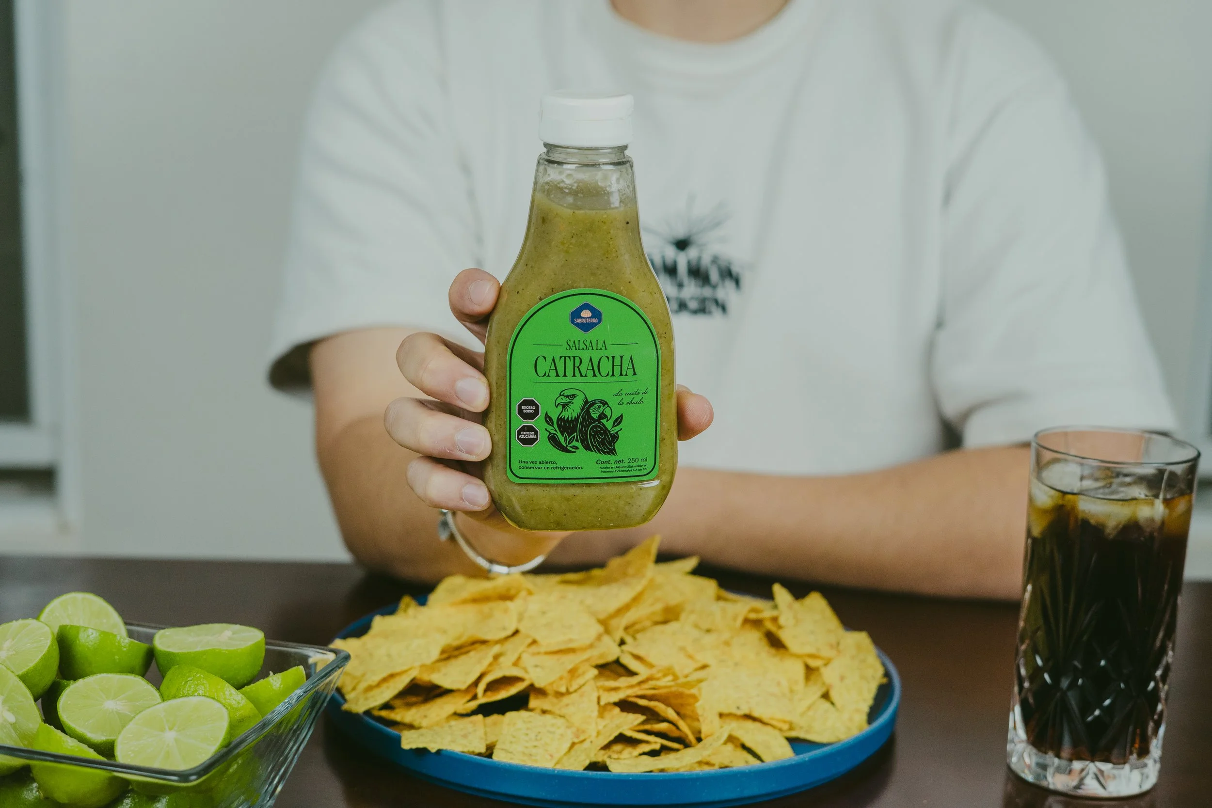 Persona sosteniendo una botella de salsa Catracha, junto a una charola de totopos, limones partidos, y un vaso con hielo y una bebida oscura.
