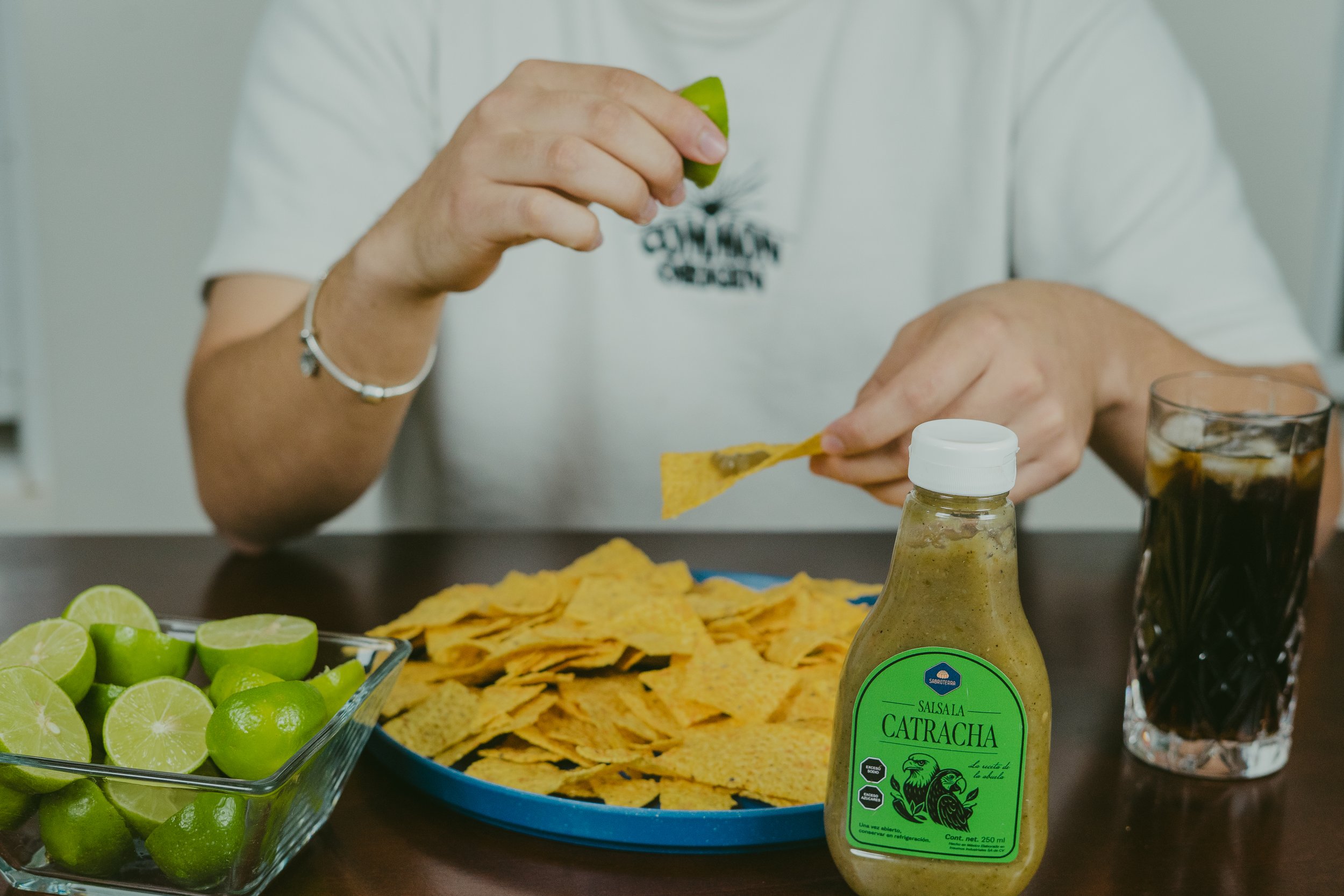 Persona sirviendo salsa cátracha sobre totopos en una mesa con limones partido, un vaso con refresco y un toque de salsa cátracha.