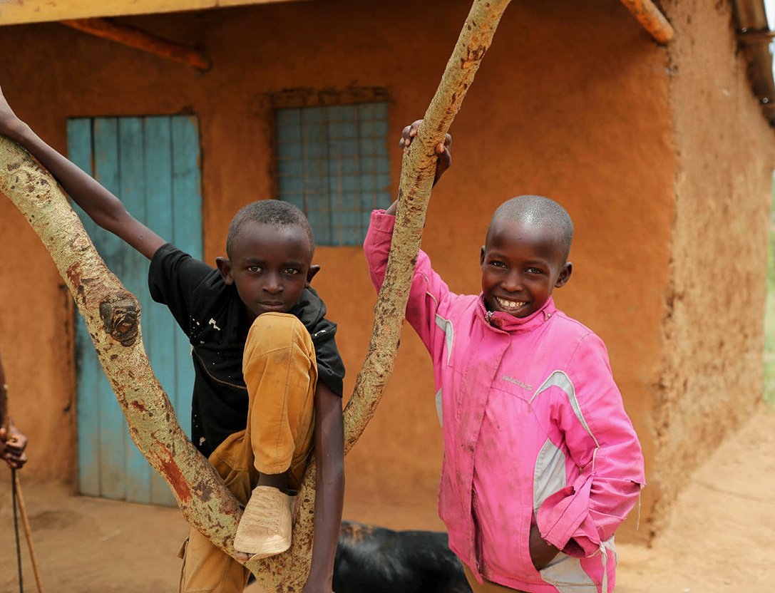 Two young boys with short hair smiling and posing with a tree in front of a mud house with a blue door in a rural setting.