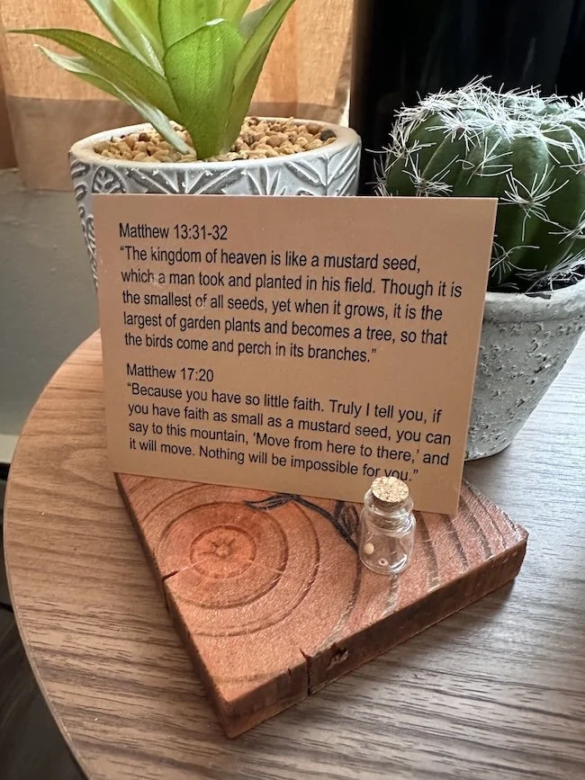 A small wooden plaque with religious quotes from Matthew 13:31-32 and Matthew 17:20, placed on a round wooden surface. Behind the plaque, there are two potted plants: one in a white decorative pot with broad green leaves, and a cactus in a gray speckled pot.