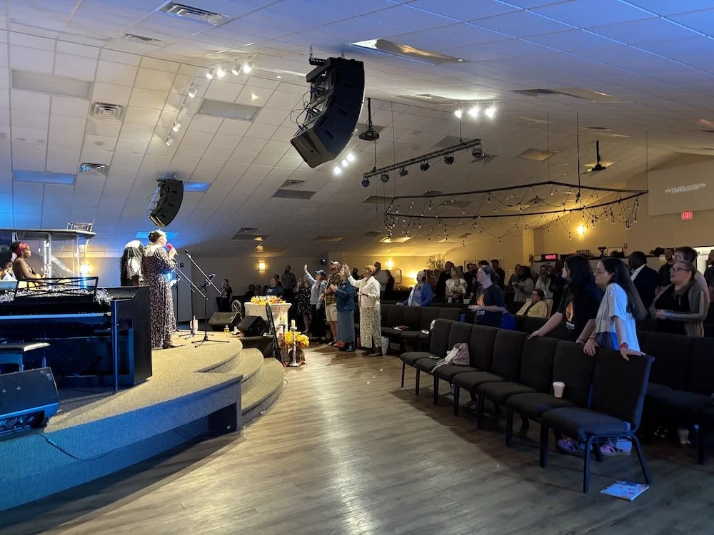 People singing and worshiping on stage in a church or religious setting, with members standing and raising hands, banners and decorative lights hanging from ceiling.
