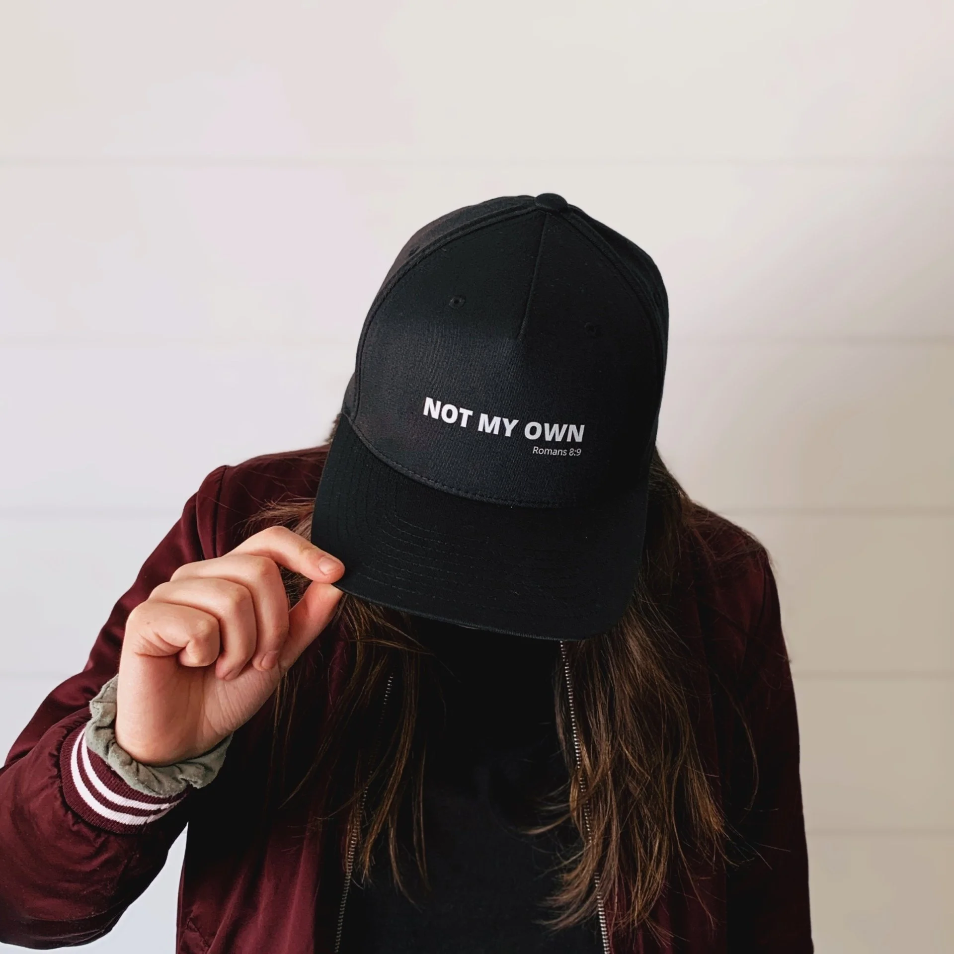 Person wearing a maroon jacket with a gray scrunchie on their wrist, holding a black cap that says 'NOT MY OWN' and references Romans 8:9.