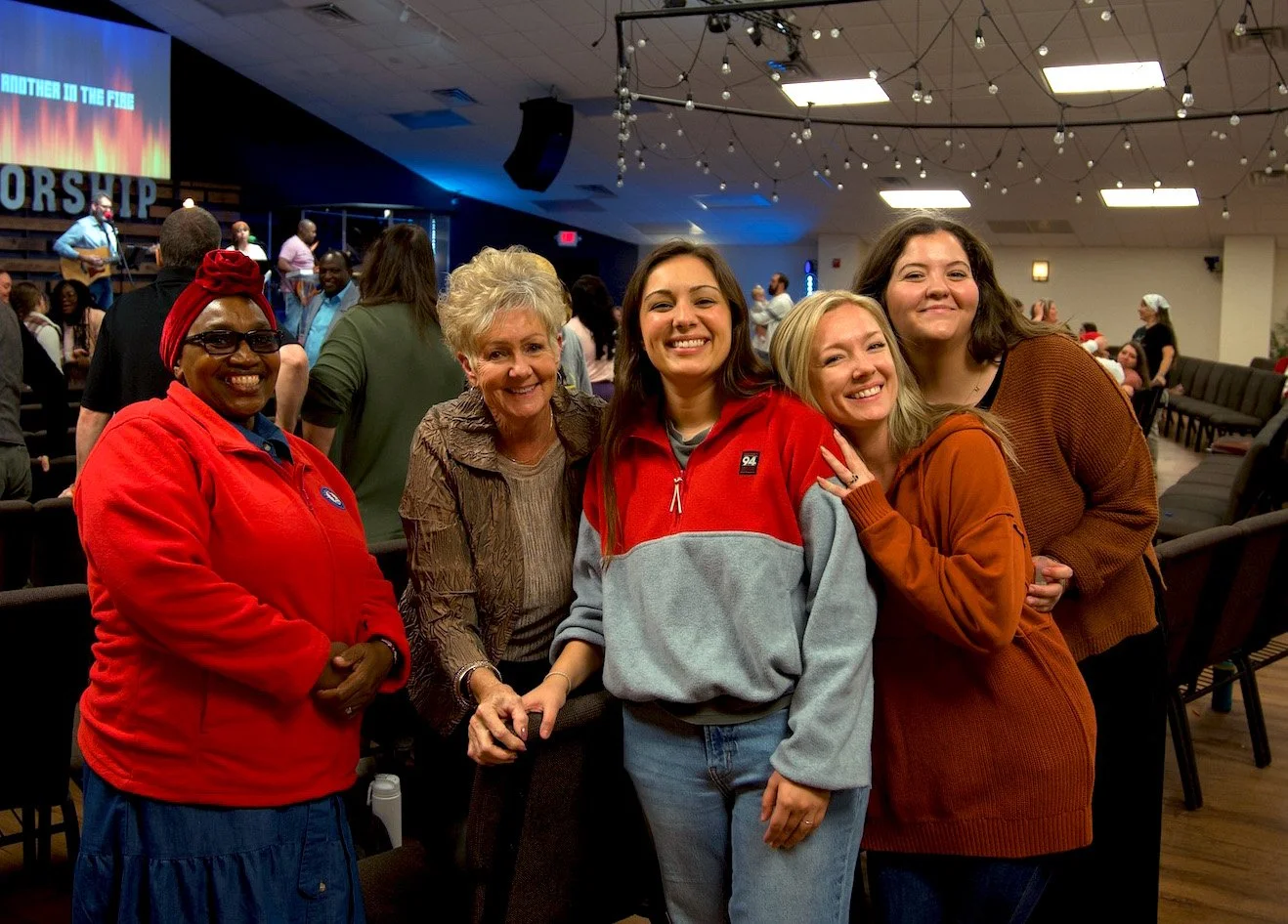 A group of five women smiling for a photo at a church or community gathering, with a stage and musicians in the background.
