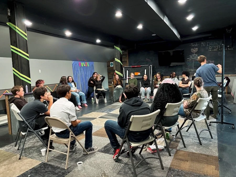 Group of people sitting in a circle in a room with black walls and ceiling, participating in a discussion or workshop, some taking notes, with a man standing and speaking to the group.