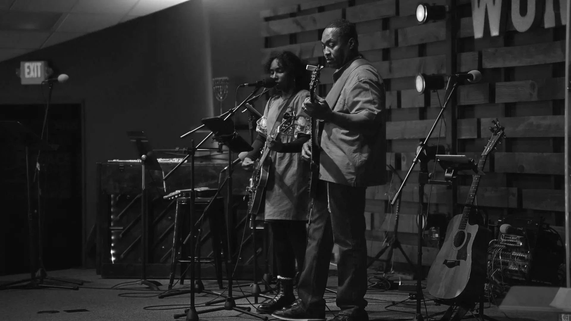 Two musicians with guitars performing on stage in a black and white photo, with a wooden-paneled wall in the background and microphones in front of them.