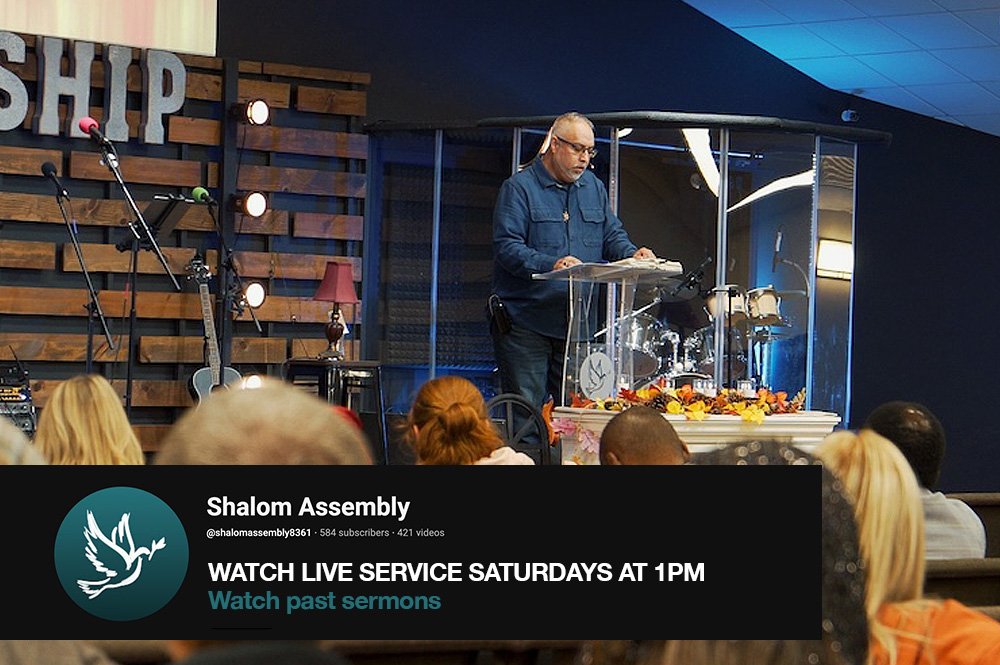 A man stands behind a transparent pulpit on a stage with a drum set and microphones, speaking to an audience in a church or assembly setting.
