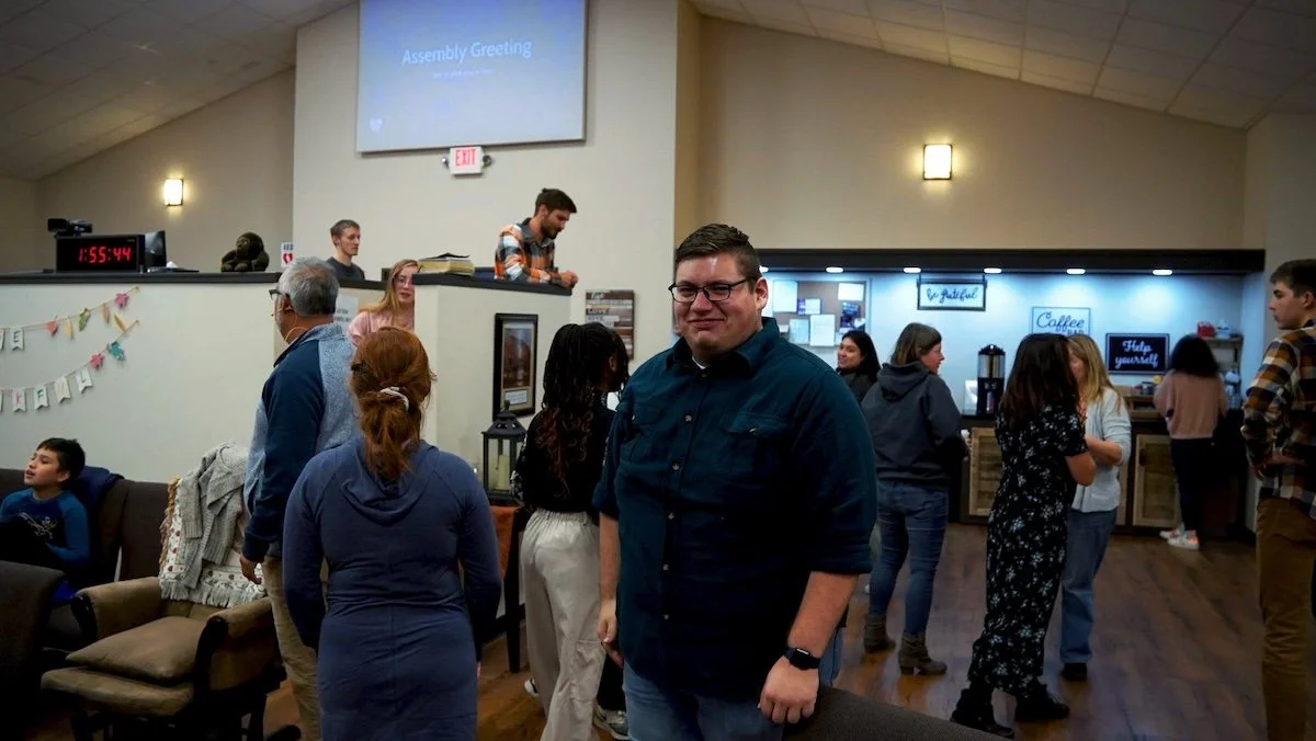 People gather in a room with a coffee bar, some socialize, others wait in line. A man in the foreground wears glasses and a dark shirt, smiling. A large screen displays 'Assembly Greeting'.