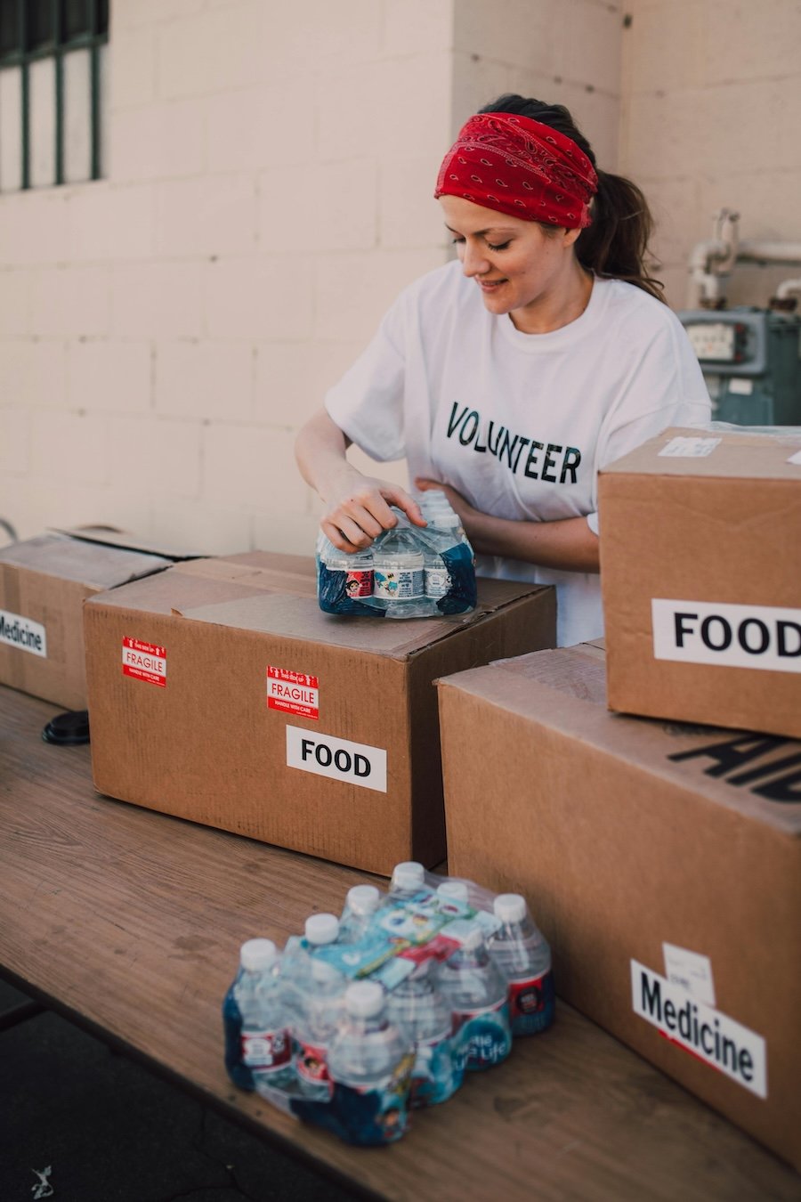 A woman in a white volunteer shirt with a red bandana on her head packing bottled water into labeled cardboard boxes for food and medicine donations.