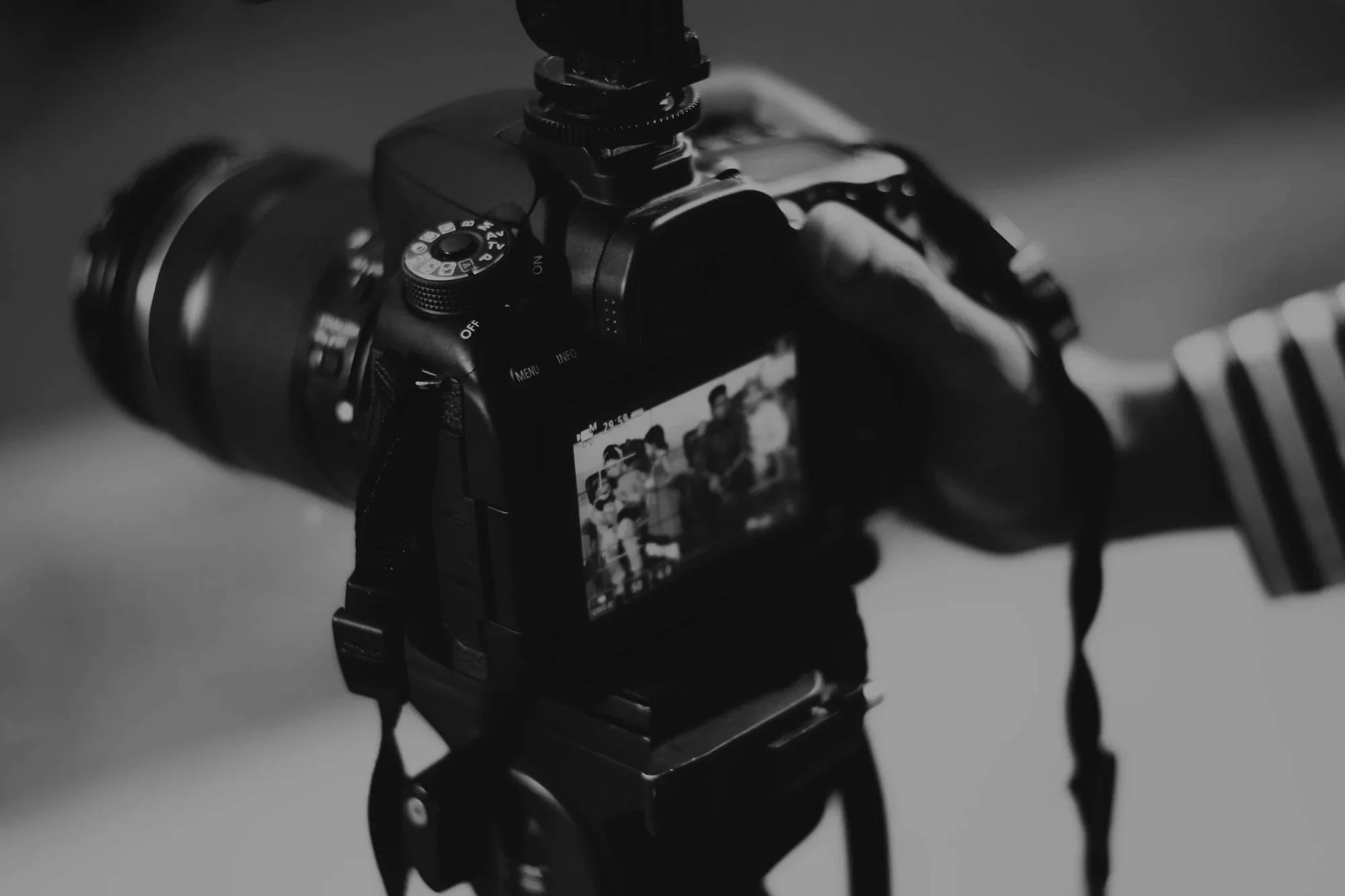 A black and white photo of a digital camera on a tripod, capturing a group of people on the camera's LCD screen.