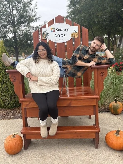 Two young women sitting on a large wooden bench outdoors with pumpkins around them. One woman is standing on the bench steps with her hands in her pockets, wearing a white sweater, black pants, and beige boots. The other is lying on top of the back of the bench, wearing a plaid shirt and jeans. Behind them is a sign that reads "Selmi's 2025 Rock Valley, IL" with autumn-themed illustrations.