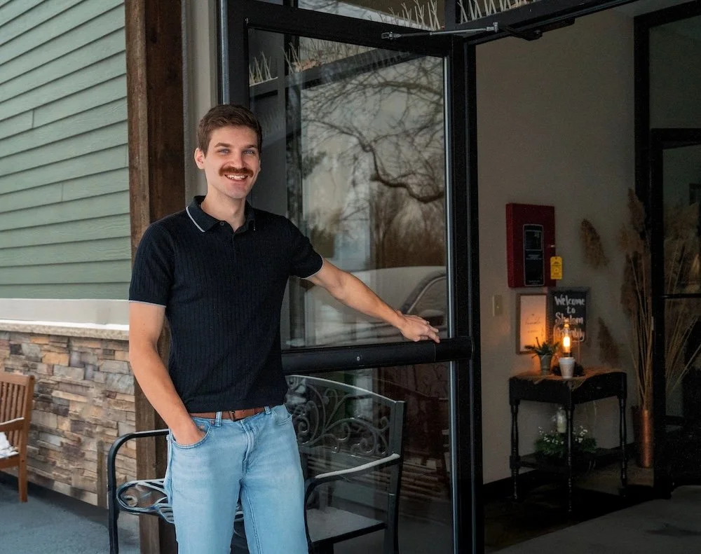 A man with a mustache, wearing a black polo shirt and light blue jeans, standing at the entrance of a building with a glass door, smiling. Contact Shalom Assembly of Yahweh In Sterling IL