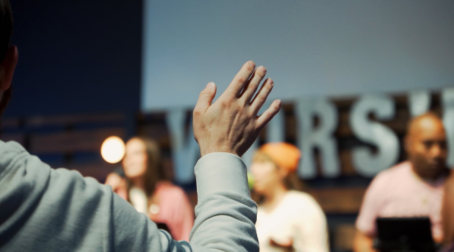 Person raising hand in a meeting or presentation with a blurred background of people and a sign that says 'MIRS'.