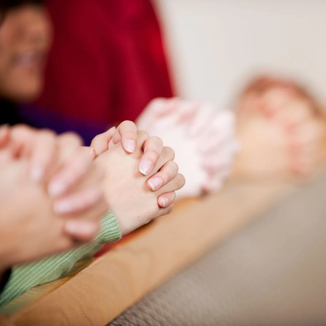 Close-up of hands clasped in prayer on a table, with a person in the background near a figure lying on a table.