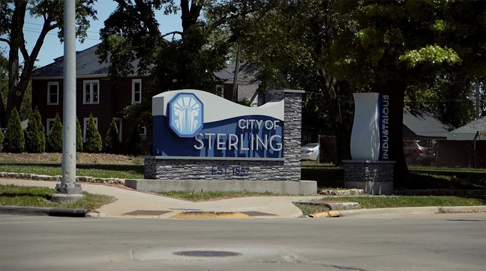 City of Sterling welcome sign with city emblem and a flag that reads 'Industrial &' in a suburban area with trees and houses in the background.