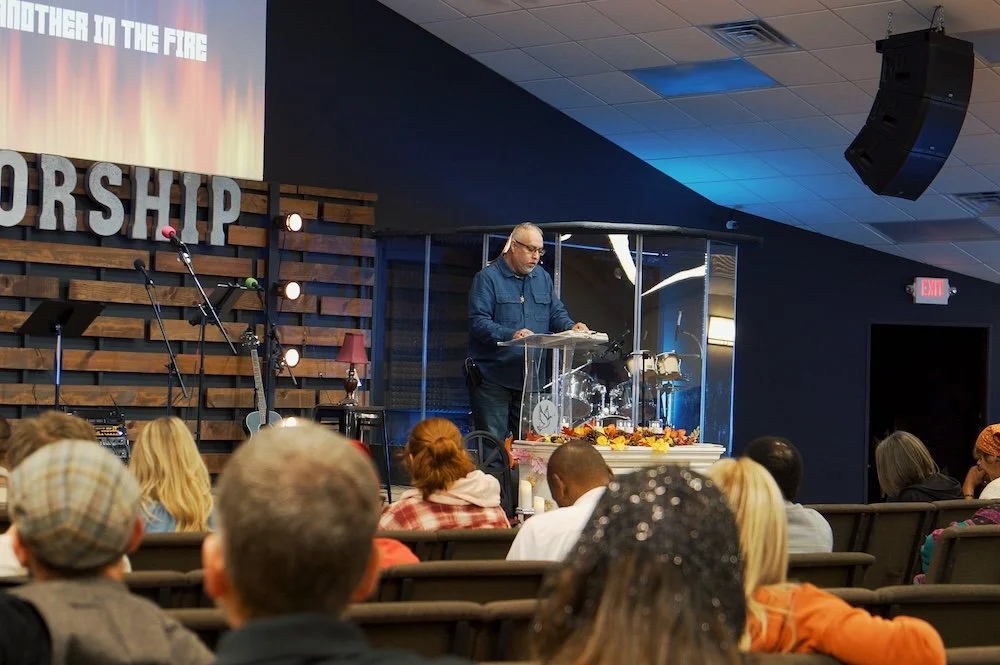 A man speaking at a church in sterling, IL. An event with an audience seated in front. The stage has a wooden backdrop with the word 'WORSHIP' displayed, and musical instruments are visible.