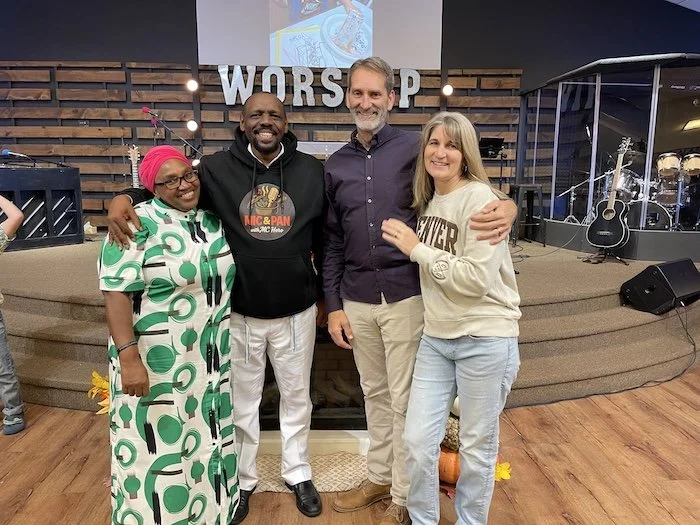 Four people standing together on a stage in a church or worship space, smiling with arms around each other, behind them the word 'WORSHIP' displayed on a wooden backdrop, musical instruments such as guitars and microphones visible.