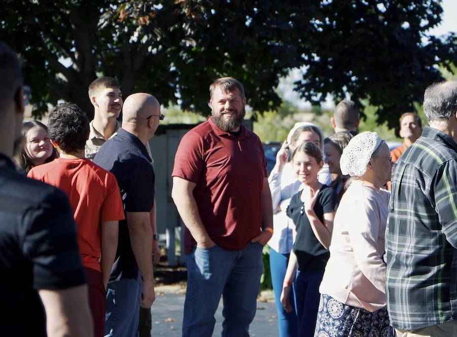 Group of people outdoors standing in line and smiling, with trees and sunlight in background.