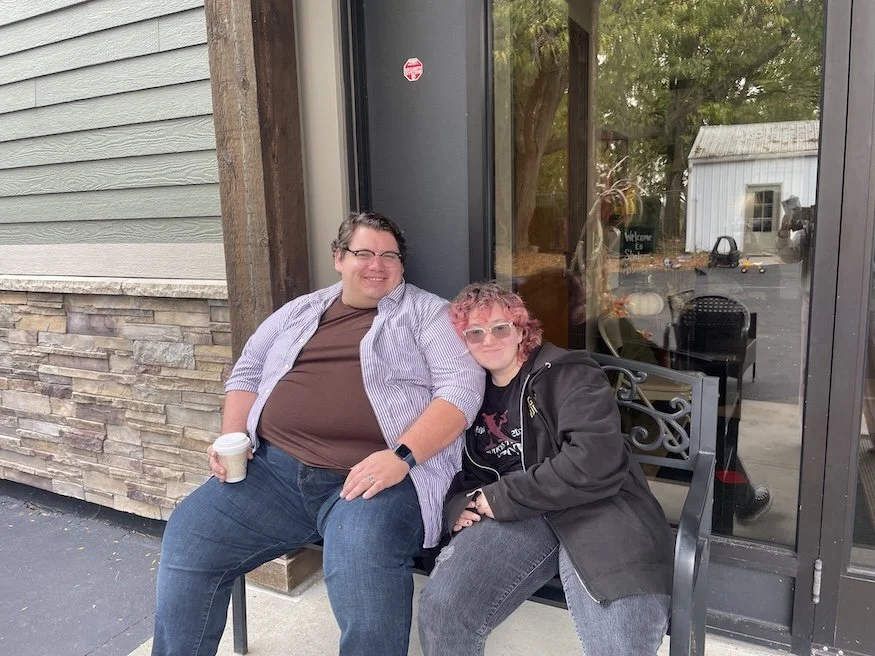 Two people sitting on a black metal bench outside a building, smiling at the camera. One person has short dark hair, glasses, and is holding a cup, and the other has pink curly hair and is wearing glasses and a black hoodie. The building has a stone and gray siding exterior with large windows reflecting a tree and a small white building in the background.