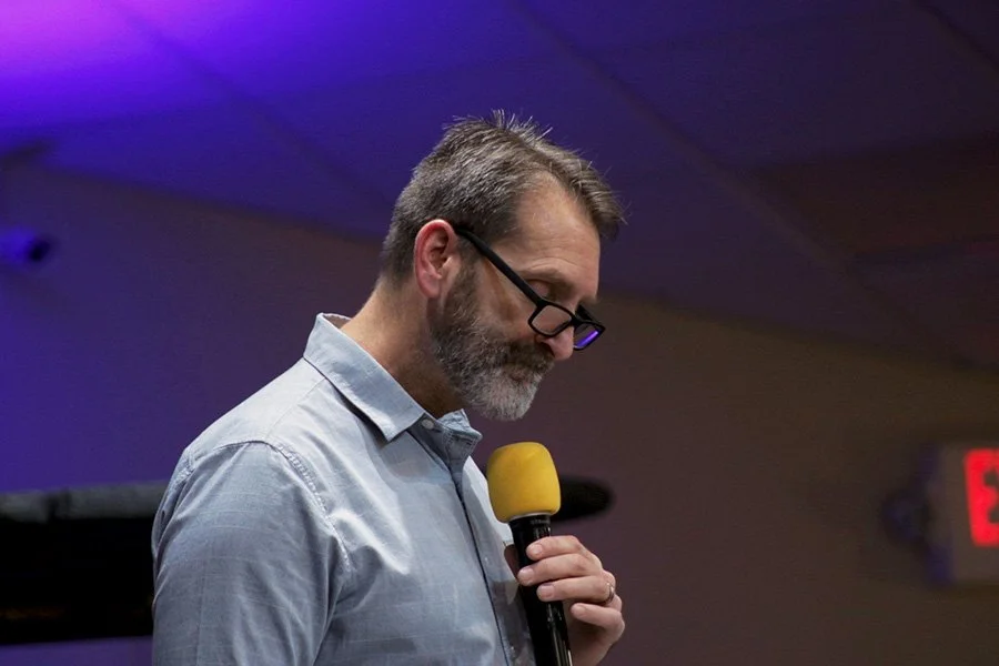 A man with glasses and a beard, wearing a light blue shirt, holding a yellow microphone during a presentation or speech in a dimly lit room.