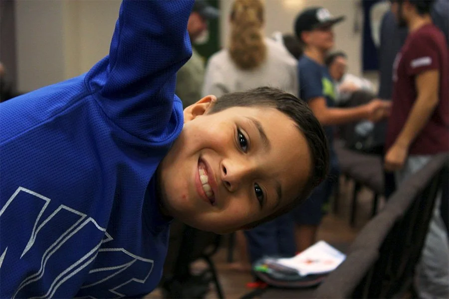 Young boy in a blue shirt smiling and leaning towards the camera, with a group of people standing and engaging in conversation in the background.