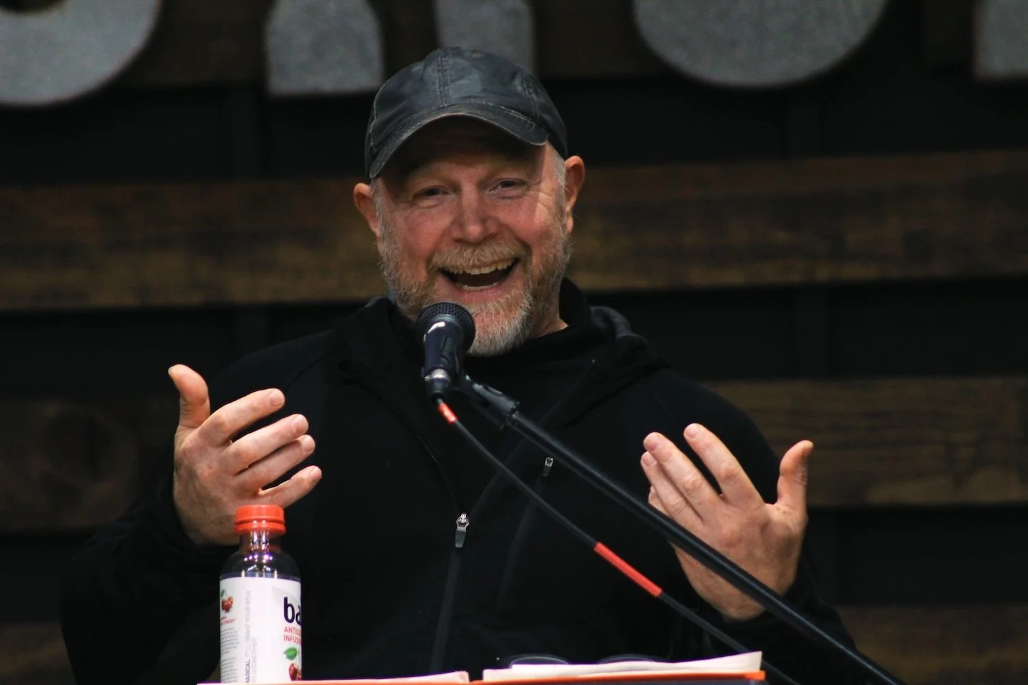 A smiling man with a beard and a cap speaking into a microphone, gesturing with his hands, with a bottle of liquid supplement on the table in front of him.