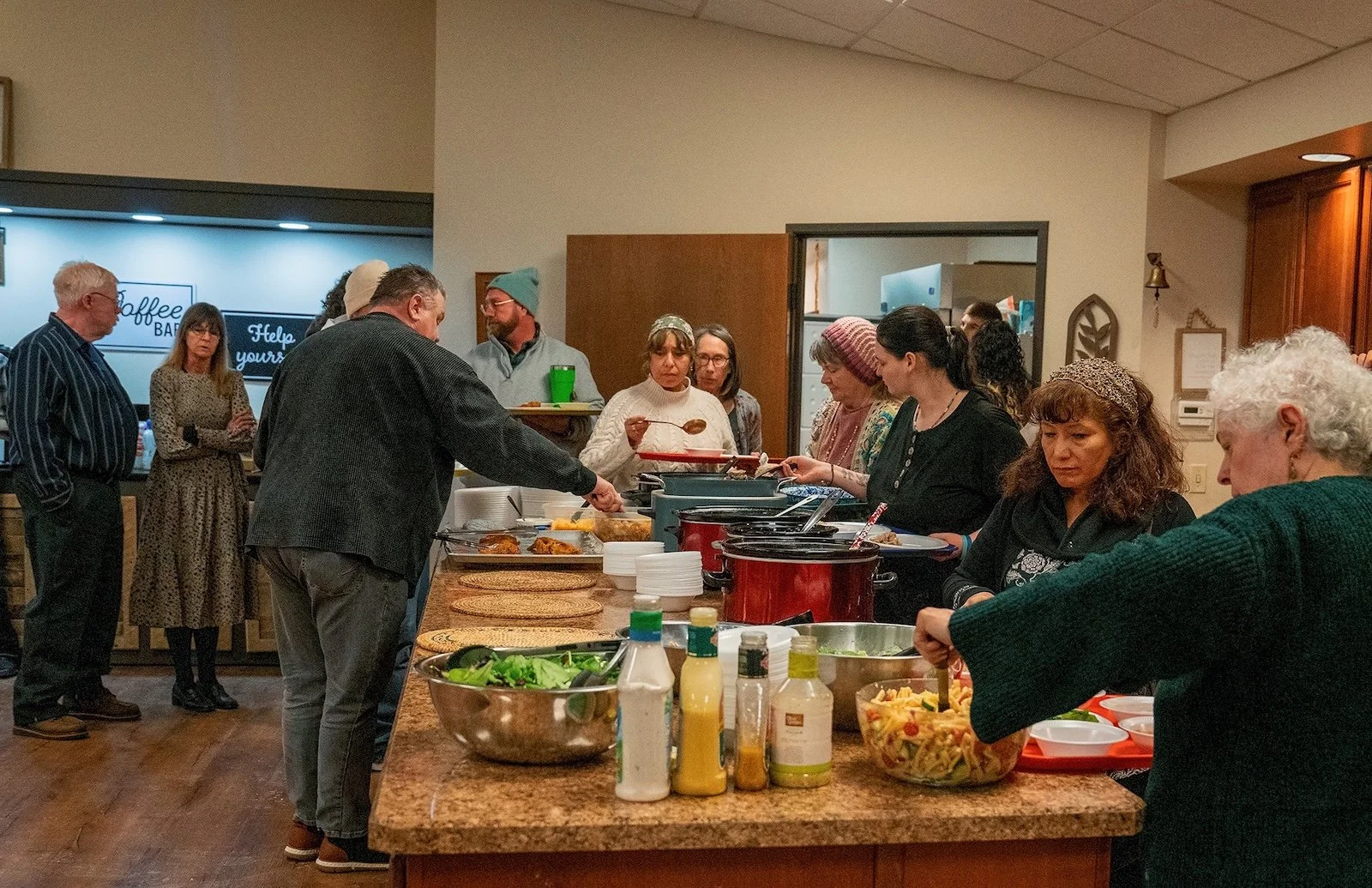 People gathered around a buffet table with food in a room, likely during a communal meal or event.