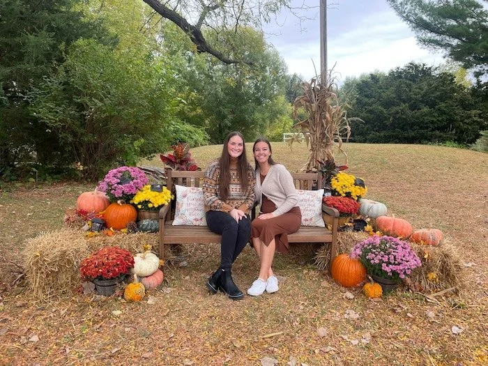 Two women sitting on a wooden bench outdoors surrounded by fall decorations, including pumpkins, flowers, and hay bales.