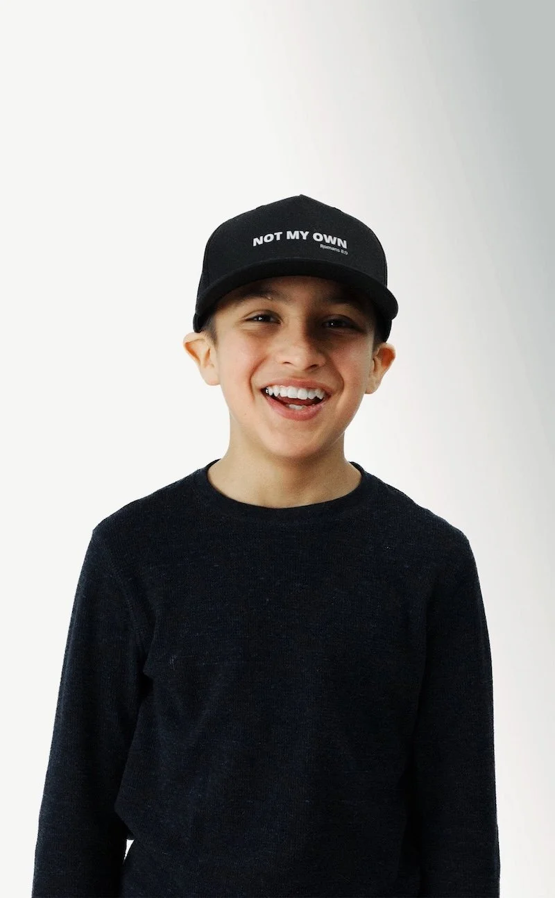 A smiling young boy wearing a black cap and black shirt, standing against a white background.