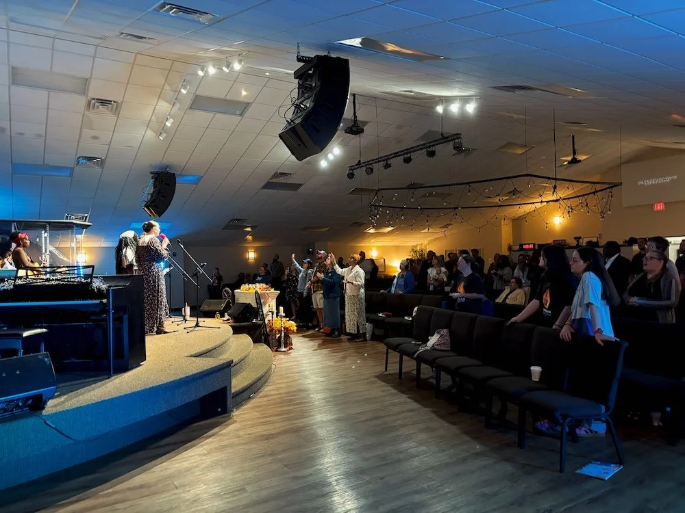 People gathered in a church auditorium listening to a woman speaking at a microphone on a stage, with a pianist nearby, during a church service or event