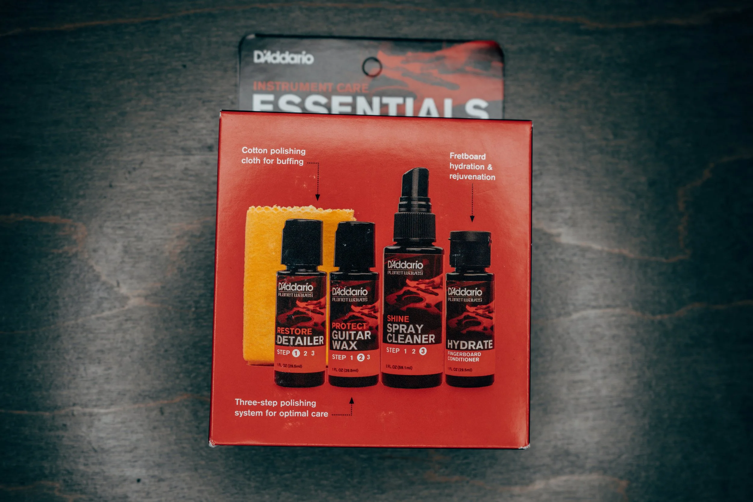 A red box containing guitar care products from D'Addario, including Restore Detailer, Protect Guitar Wax, Spray Cleaner, and Hydrate fingerboard conditioner, with a yellow polishing cloth depicted on the packaging.