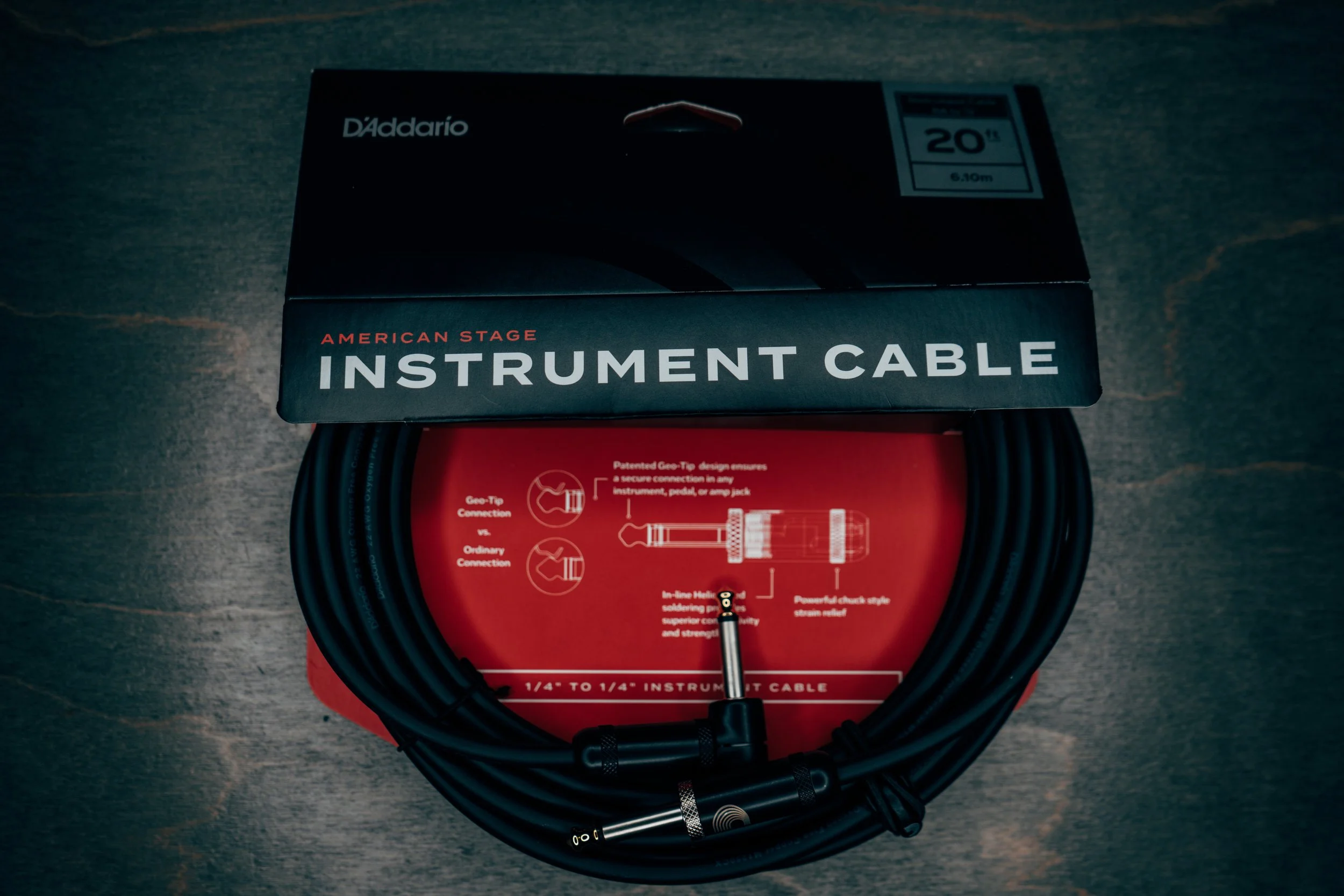 A box of D'Addario American Stage instrument cables with coiled black cable and a 1/4 inch connector on the end, resting on a wooden surface.