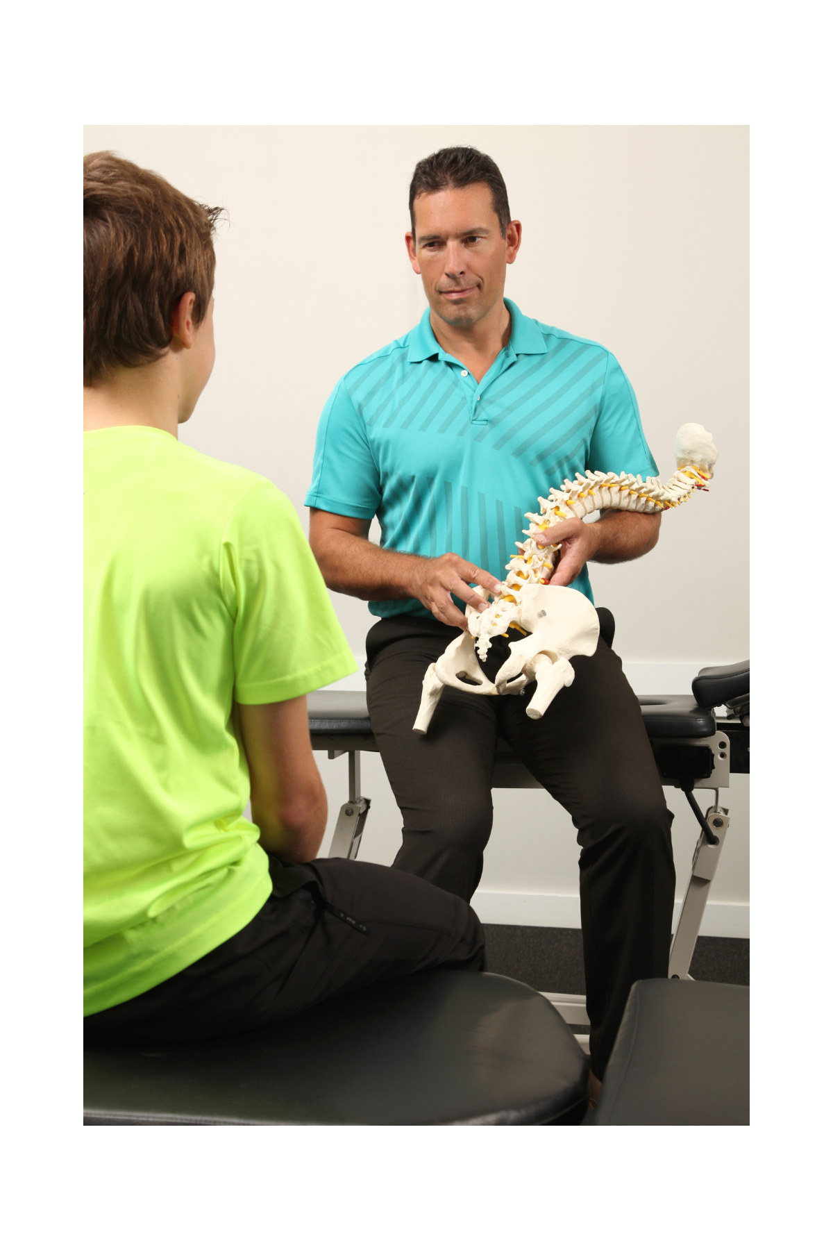 A man in a turquoise shirt is demonstrating a model of a human spinal column and pelvis to a young person in a neon green shirt in a clinical setting.