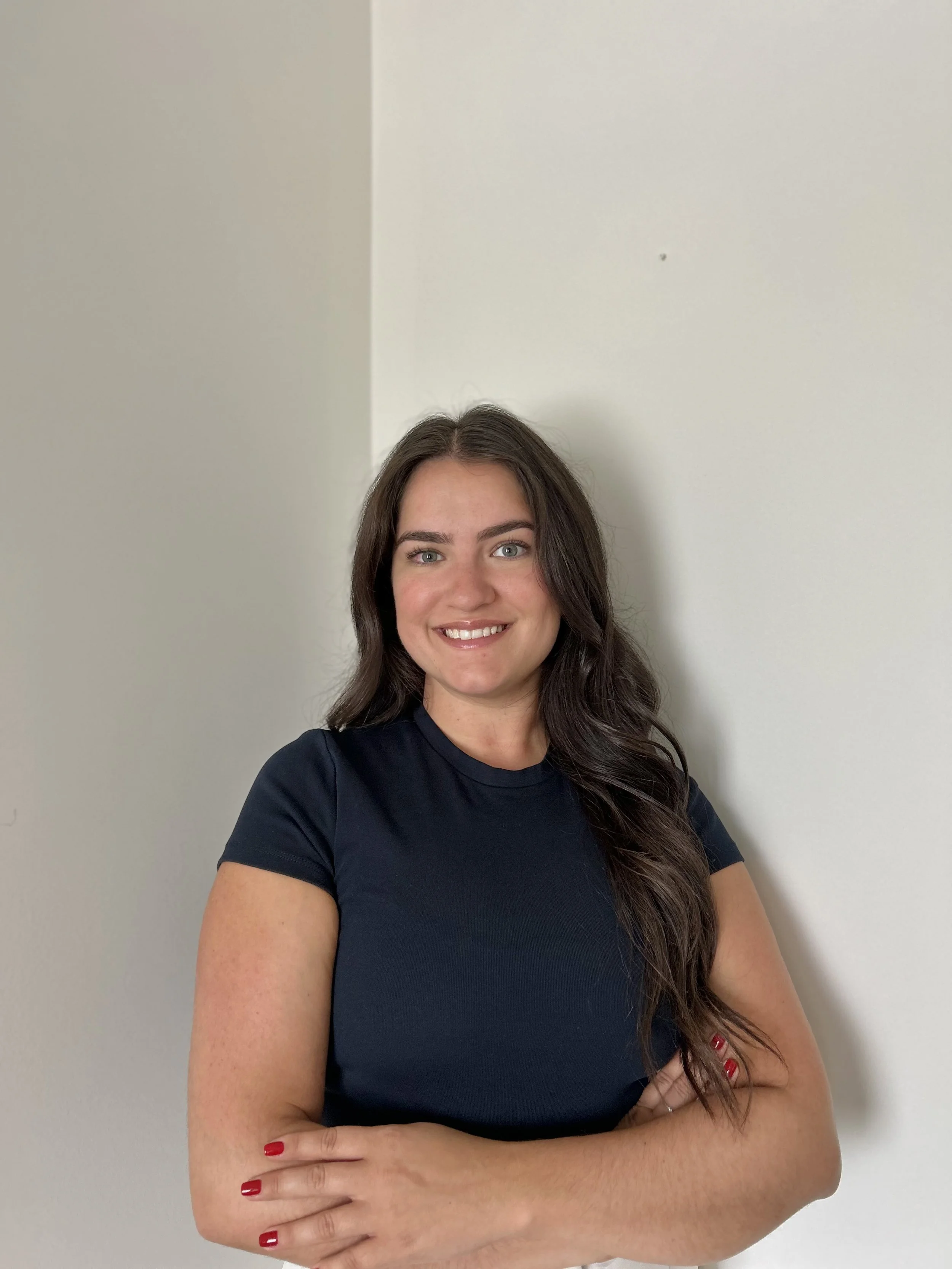 A smiling woman with short light brown hair and blue eyes, wearing a dark blue polo shirt and black pants, standing with arms crossed in front of a plain light-colored wall.