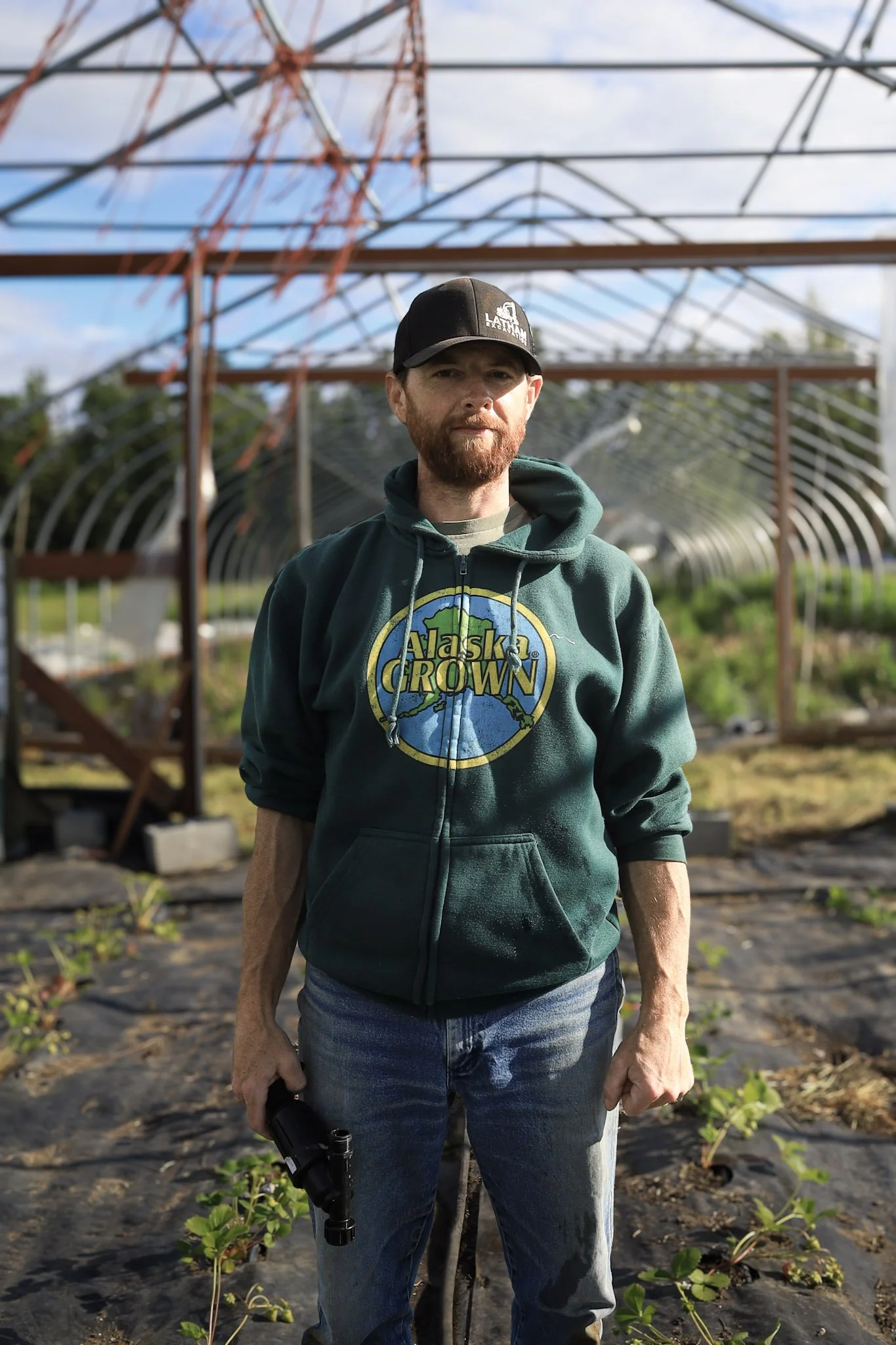 A man standing in a greenhouse, holding a small gardening tool, wearing a green hoodie with 'Alaska Grown' logo, a black cap, and jeans, with plants and a metal arched structure in the background.