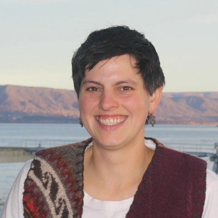 A woman with short dark hair smiling, wearing earrings and a patterned vest, outdoors with mountains and water in the background.