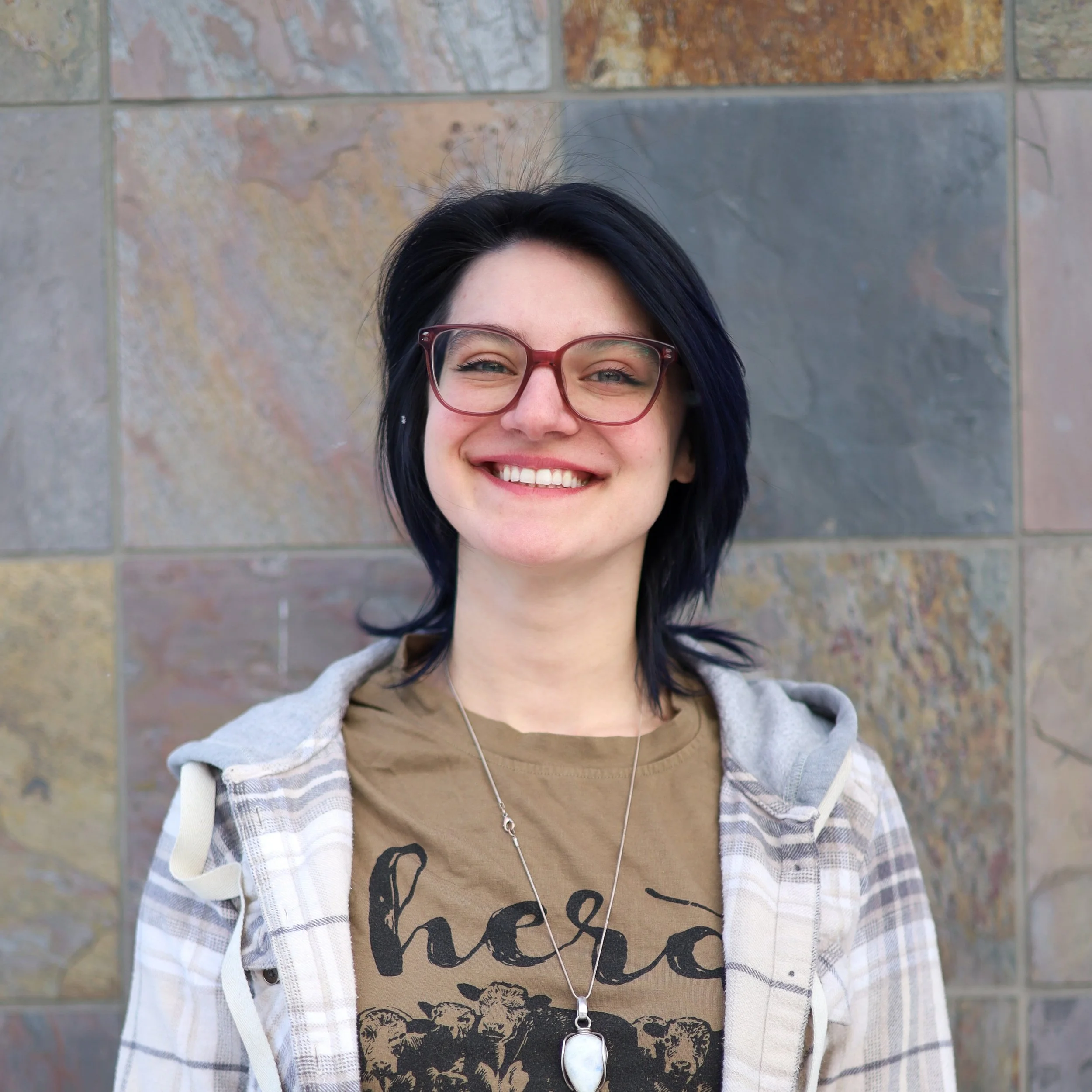 A young woman with short dark hair, glasses, a brown graphic t-shirt, and a light-colored plaid jacket, smiling in front of a tiled wall.