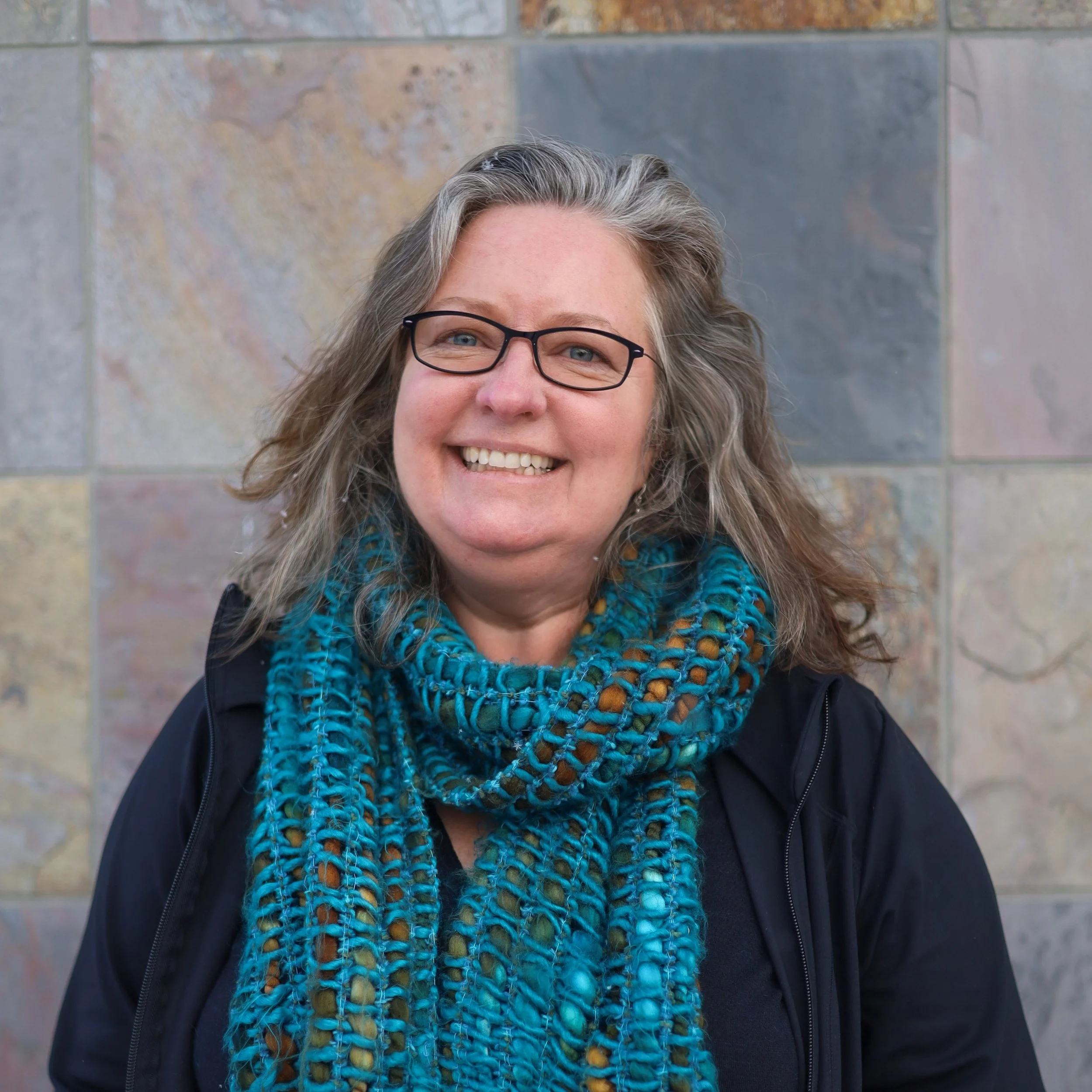 A smiling middle-aged woman with glasses, long wavy gray hair, wearing a blue patterned scarf, black jacket, standing in front of a multicolored stone wall.